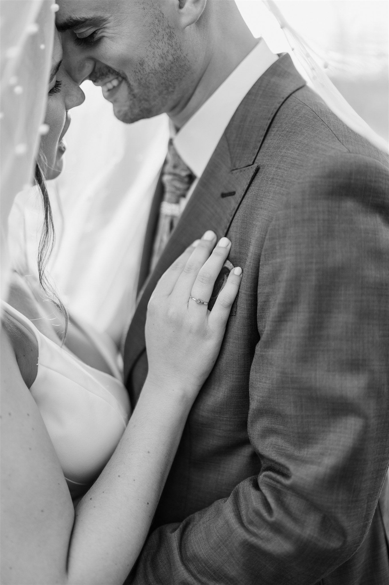 Black and white photo by Portugal Wedding Photographer of a bride and groom embracing, smiling under a veil, showing her engagement ring.