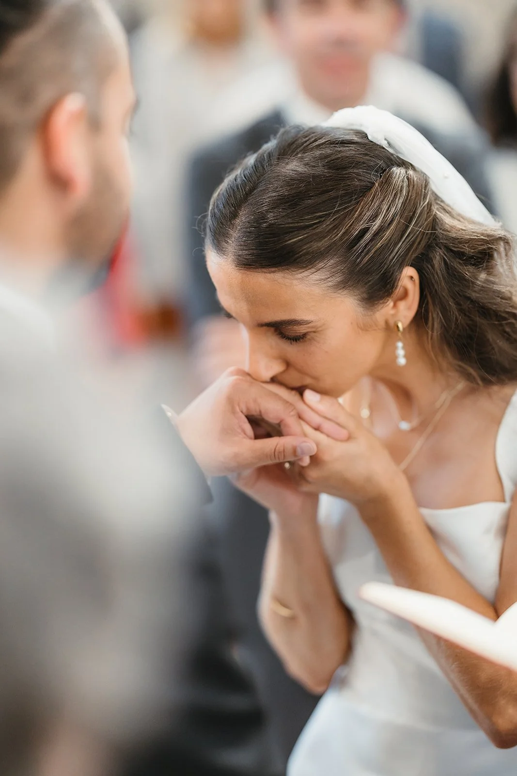 Portugal Wedding Photographer captures bride in white dress gently kissing groom’s hand during ceremony, guests blurred in background.