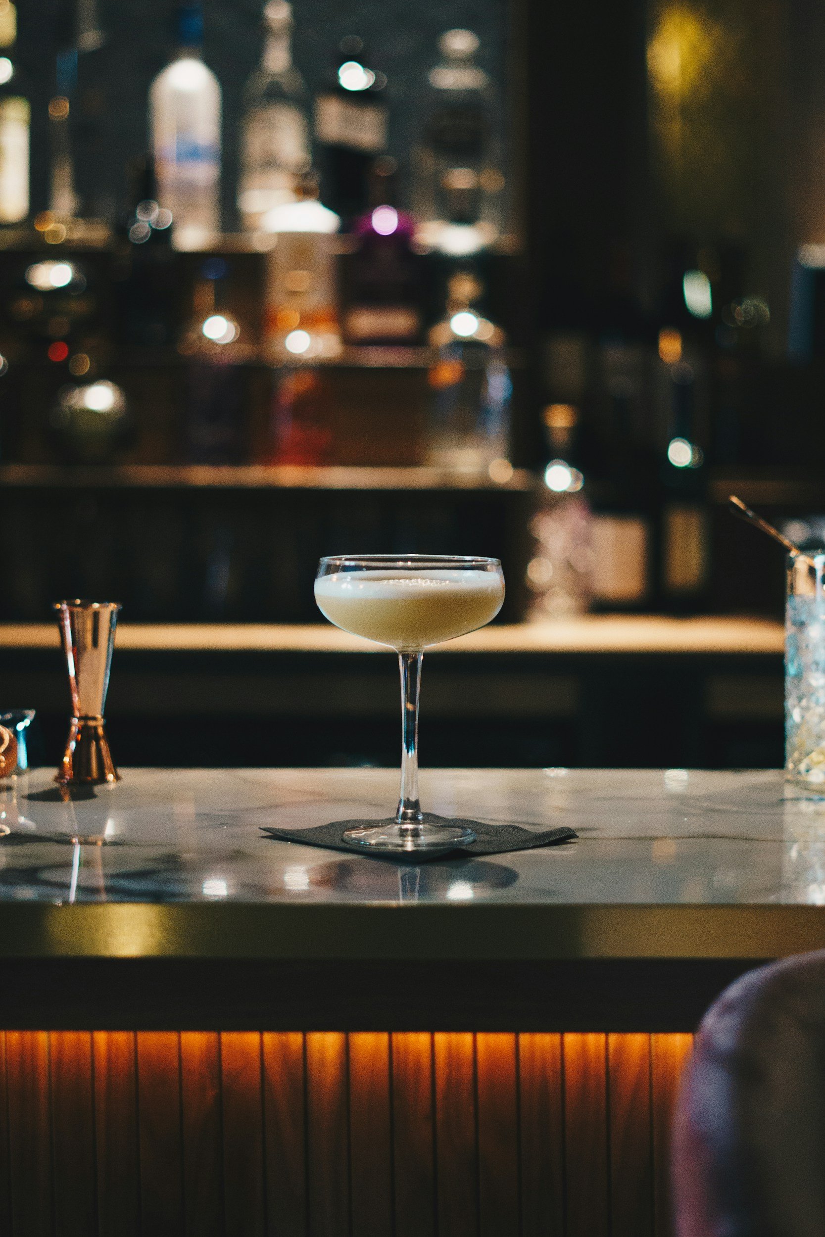A cocktail in a coupe glass on a marble bar counter with bar tools and liquor bottles in the background.
