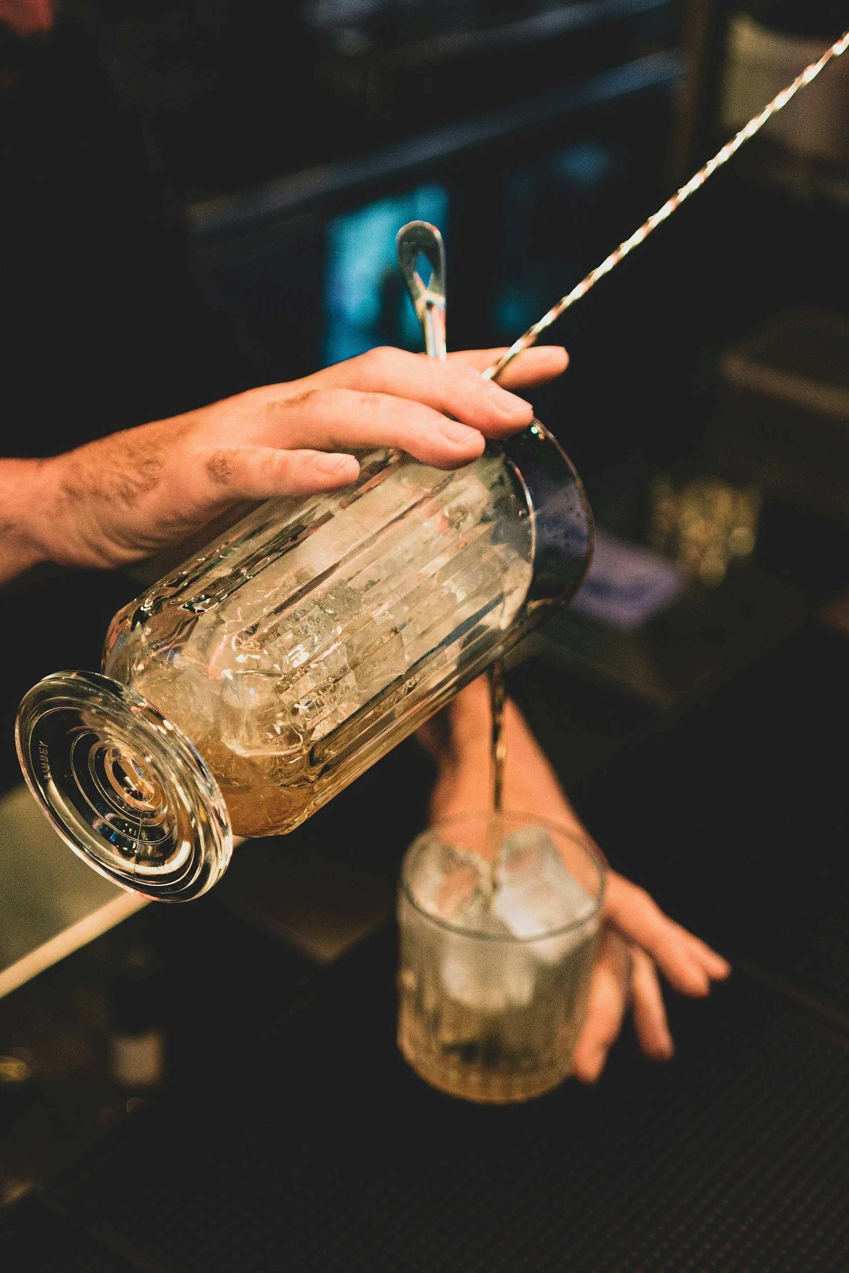 A bartender pours a drink with ice from a glass shaker into a glass with ice. The background is dark and slightly blurred.