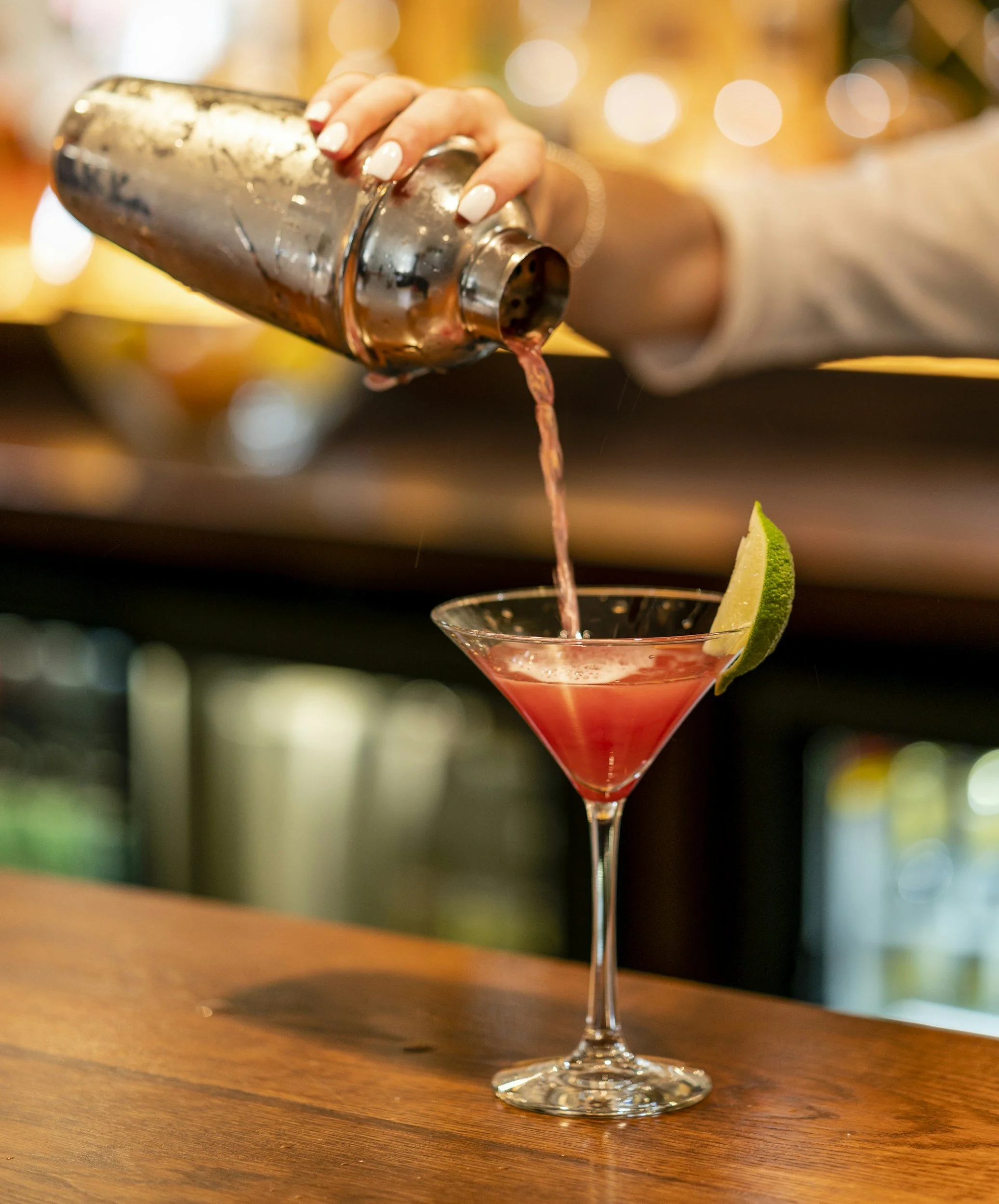 A bartender pouring a pink cocktail from a metal shaker into a martini glass garnished with a lime wedge, on a wooden bar counter.