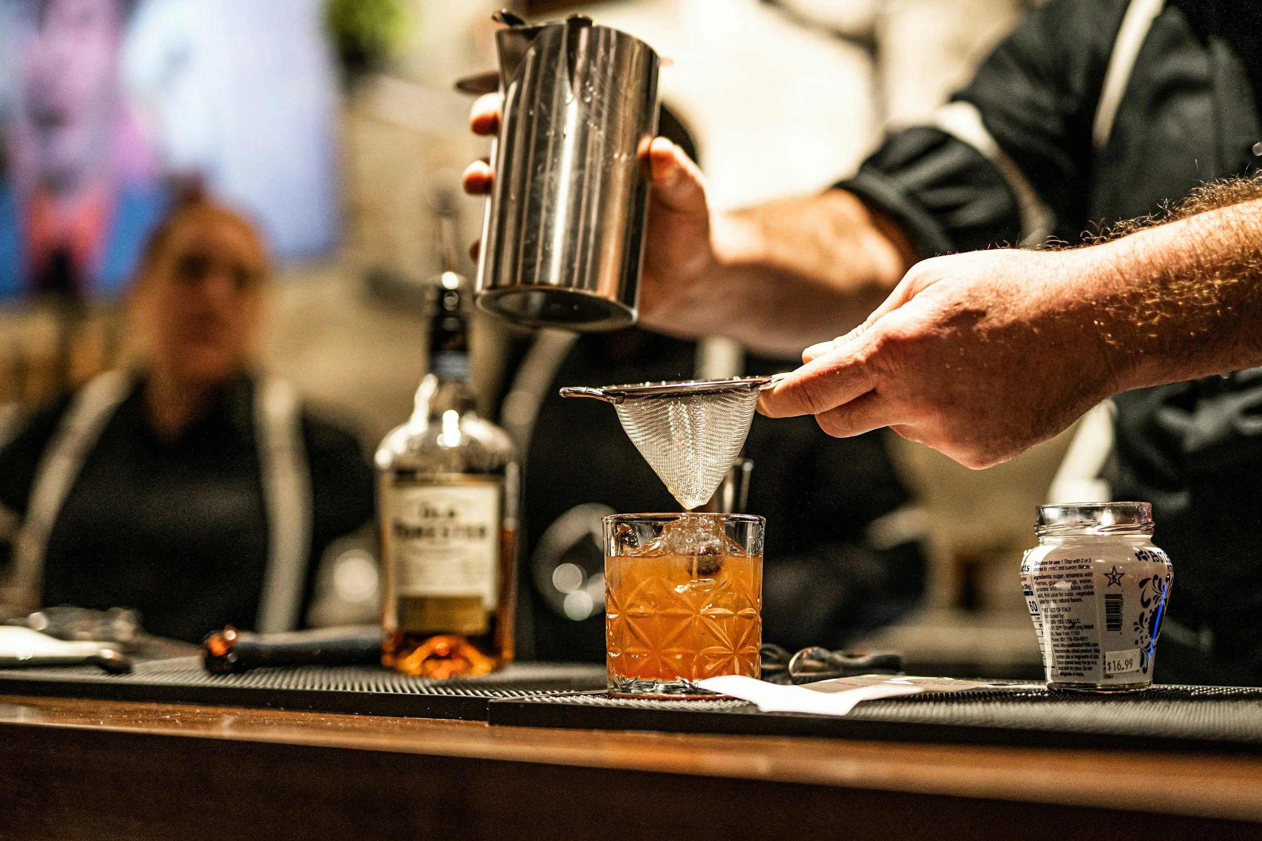 A bartender using a fine mesh strainer to pour a cocktail into a glass with a large ice cube. The scene is set at a bar with a bottle of liquor and a container of cream nearby, and a blurred woman watching in the background.