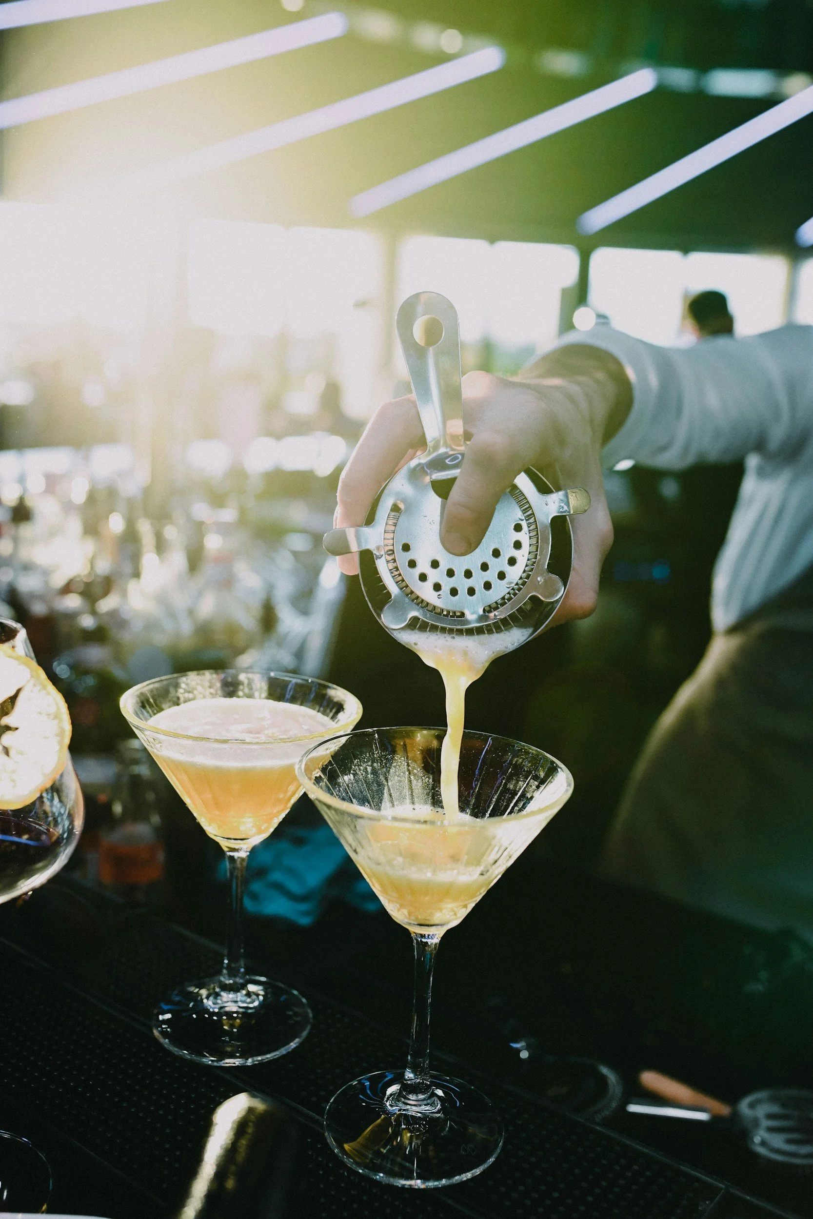 A bartender pouring a cocktail from a strainer into a martini glass at an outdoor bar with sunlight streaming in.