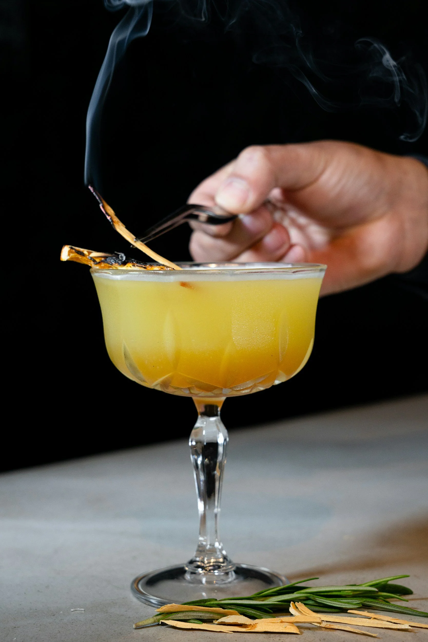 Hands holding a cocktail glass with a yellow drink, garnished with a lemon peel, as smoke rises from a flame nearby, with rosemary and toasted almonds on the table.