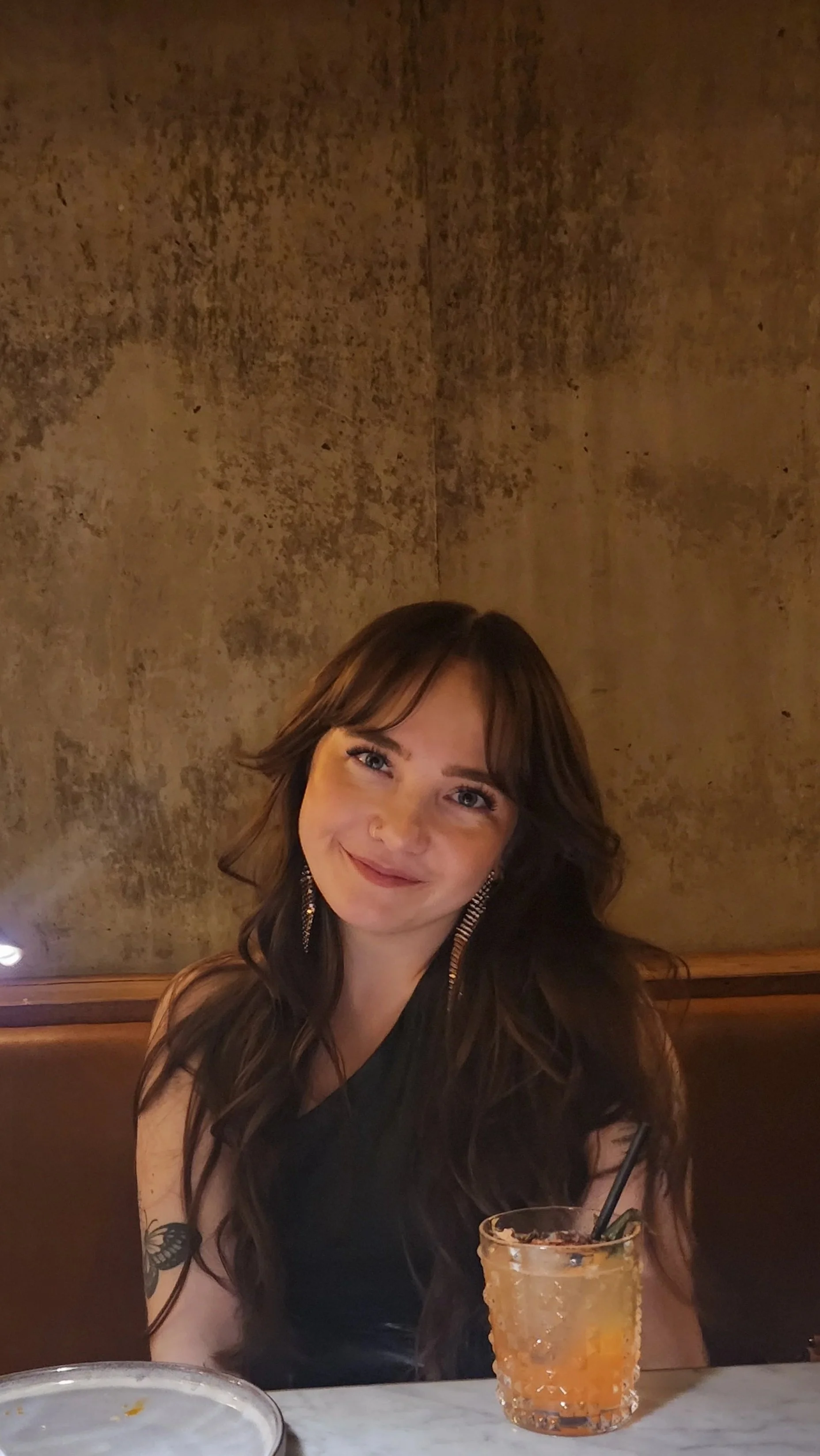 A young woman with long brown hair and earrings, smiling while sitting at a restaurant table, with a drink in a glass in front of her.