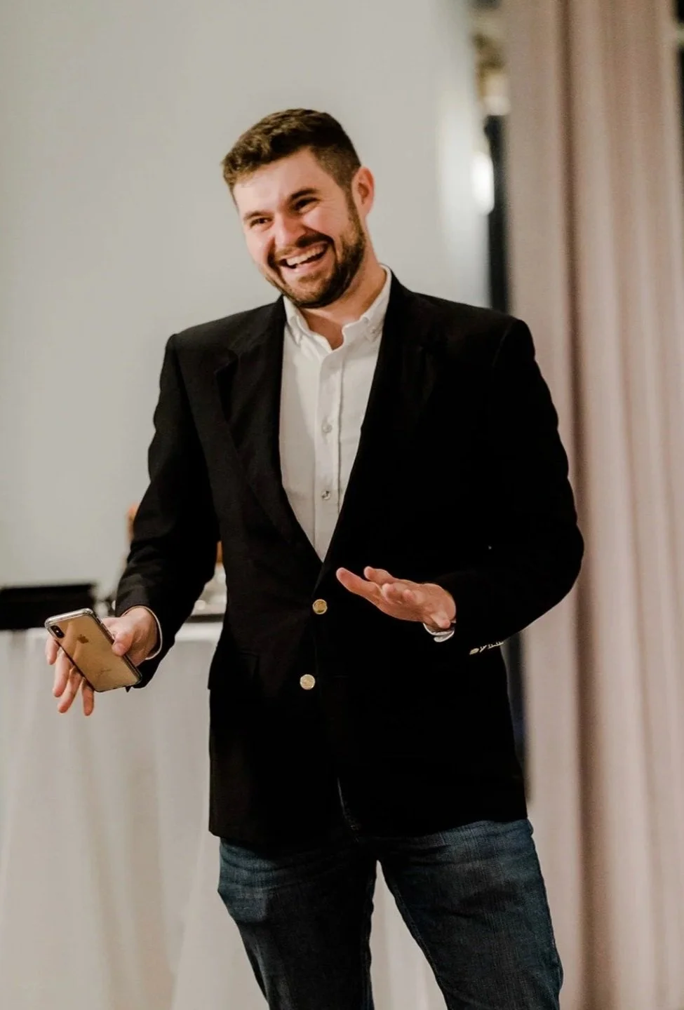 A man with a beard and short hair, dressed in a black blazer and white shirt, smiling and gesturing while holding a smartphone in his left hand in a room with beige curtains.