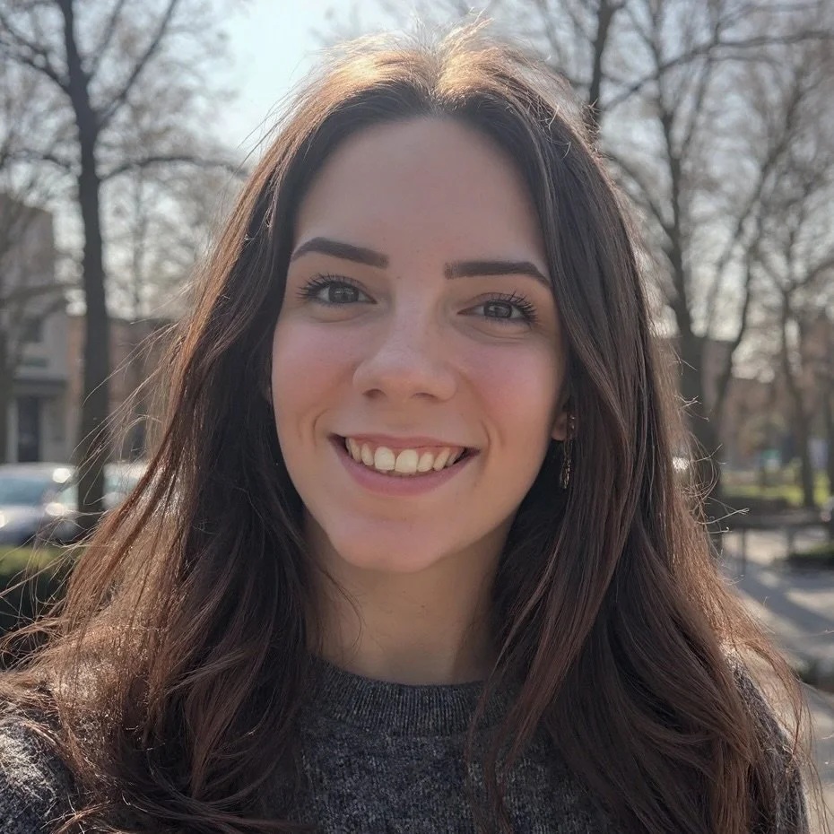 A young woman with long dark brown hair smiling outside on a sunny day, with trees and buildings in the background.