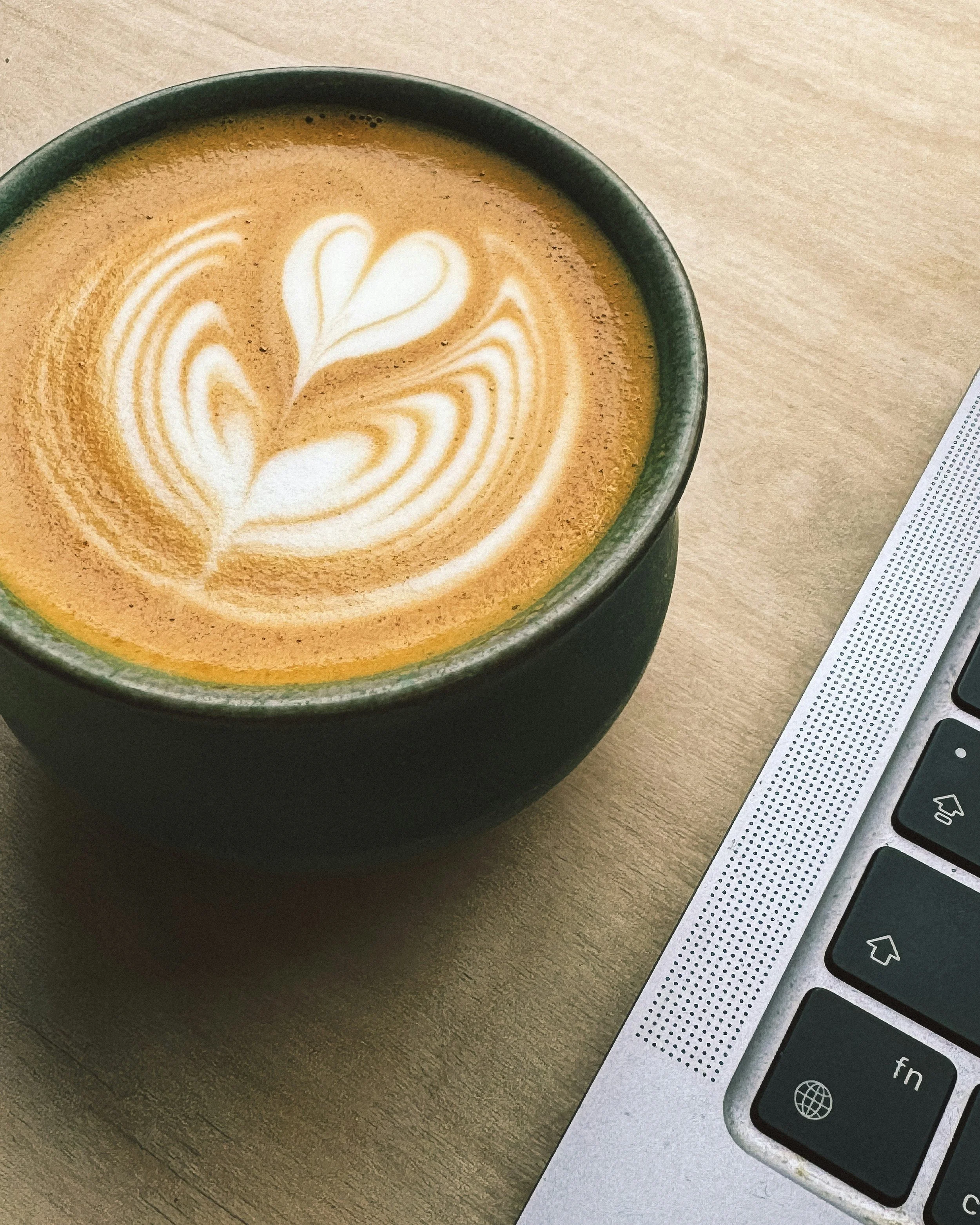 A cup of coffee with decorative foam art on a wooden table next to a laptop keyboard.