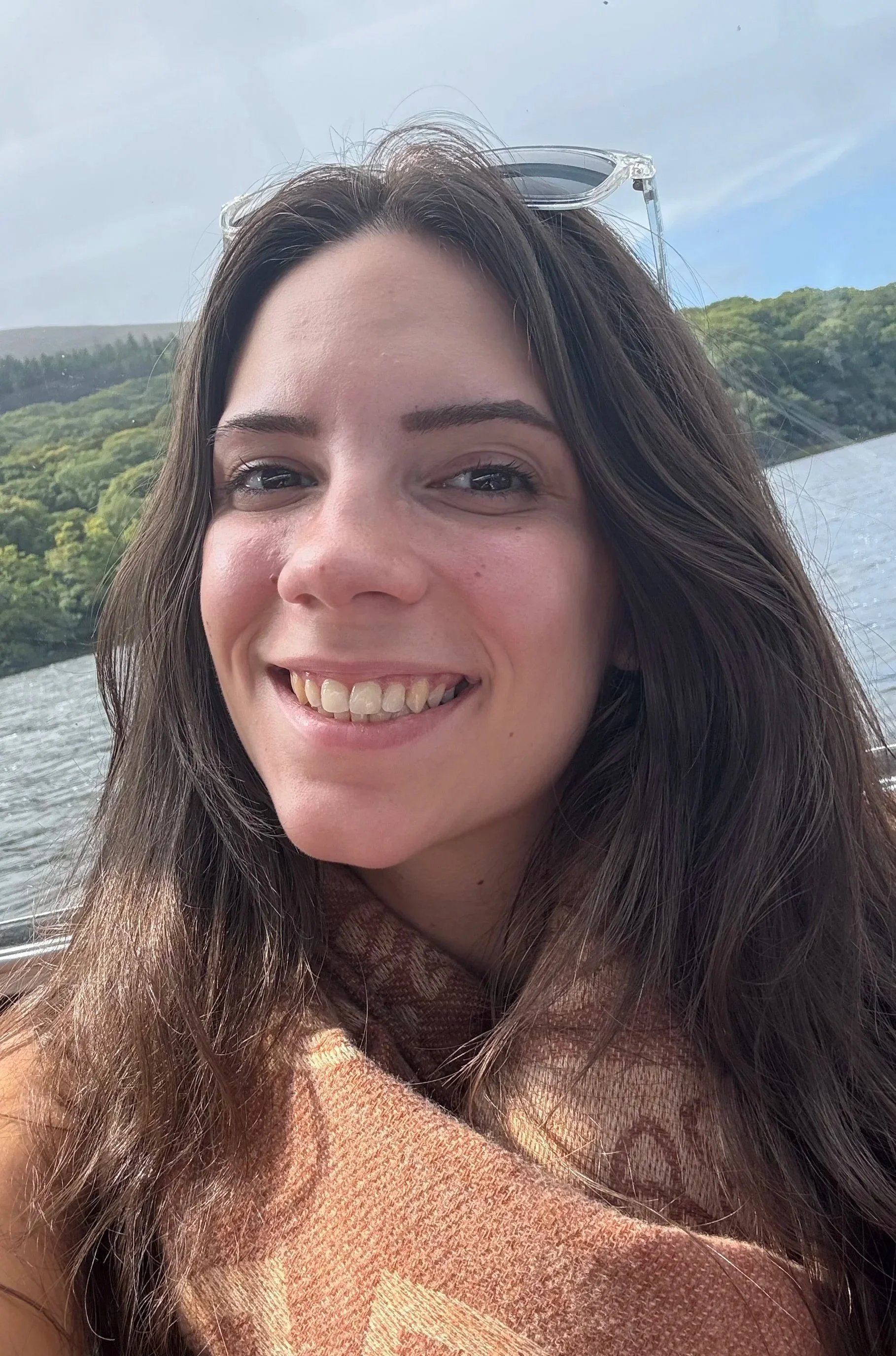 A young woman with long brown hair smiling outdoors near a body of water, with green hills and a cloudy sky in the background, wearing sunglasses pushed up on her head and a scarf around her neck.