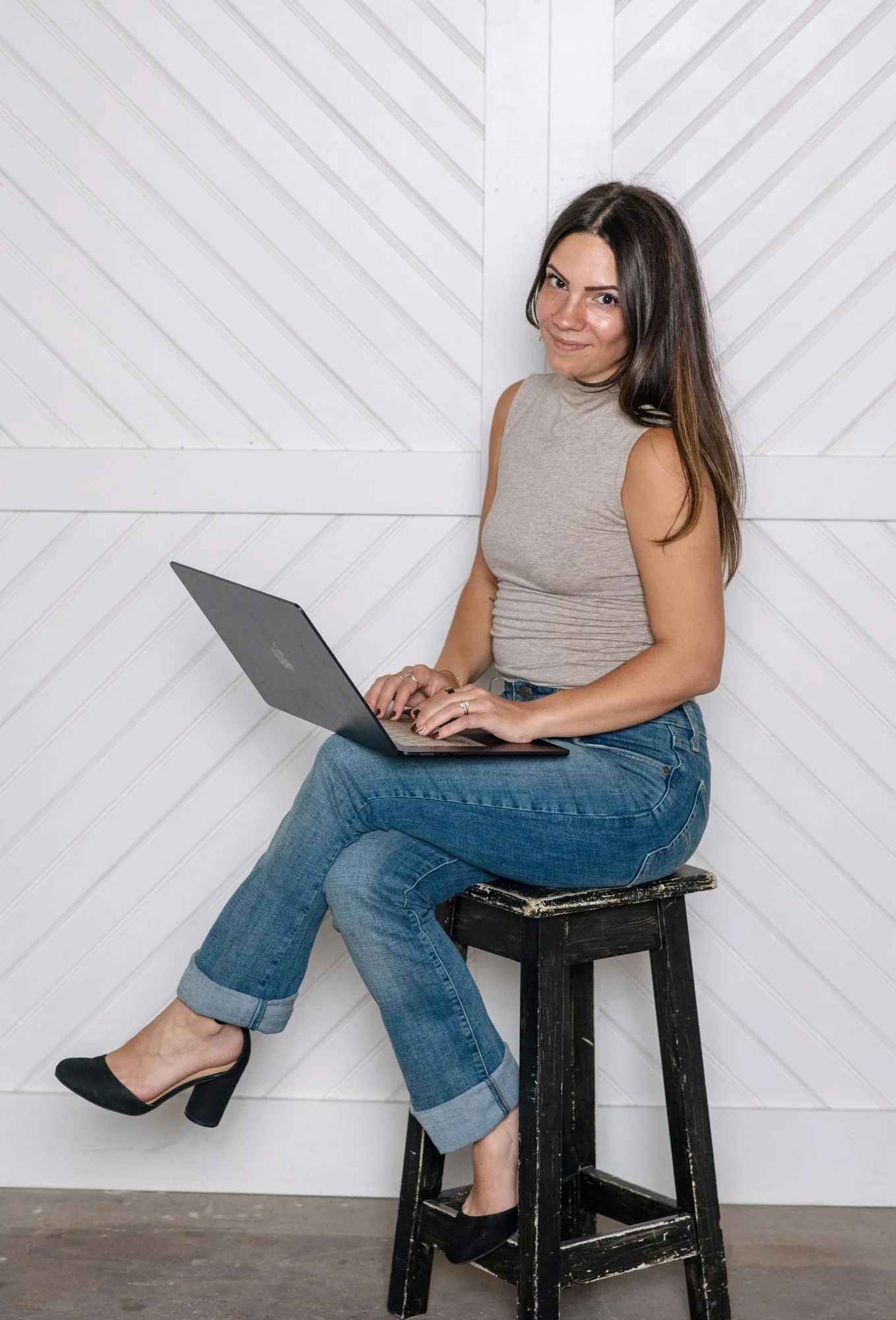 Sky sitting on a stool against a white wall, smiling with her laptop on her lap