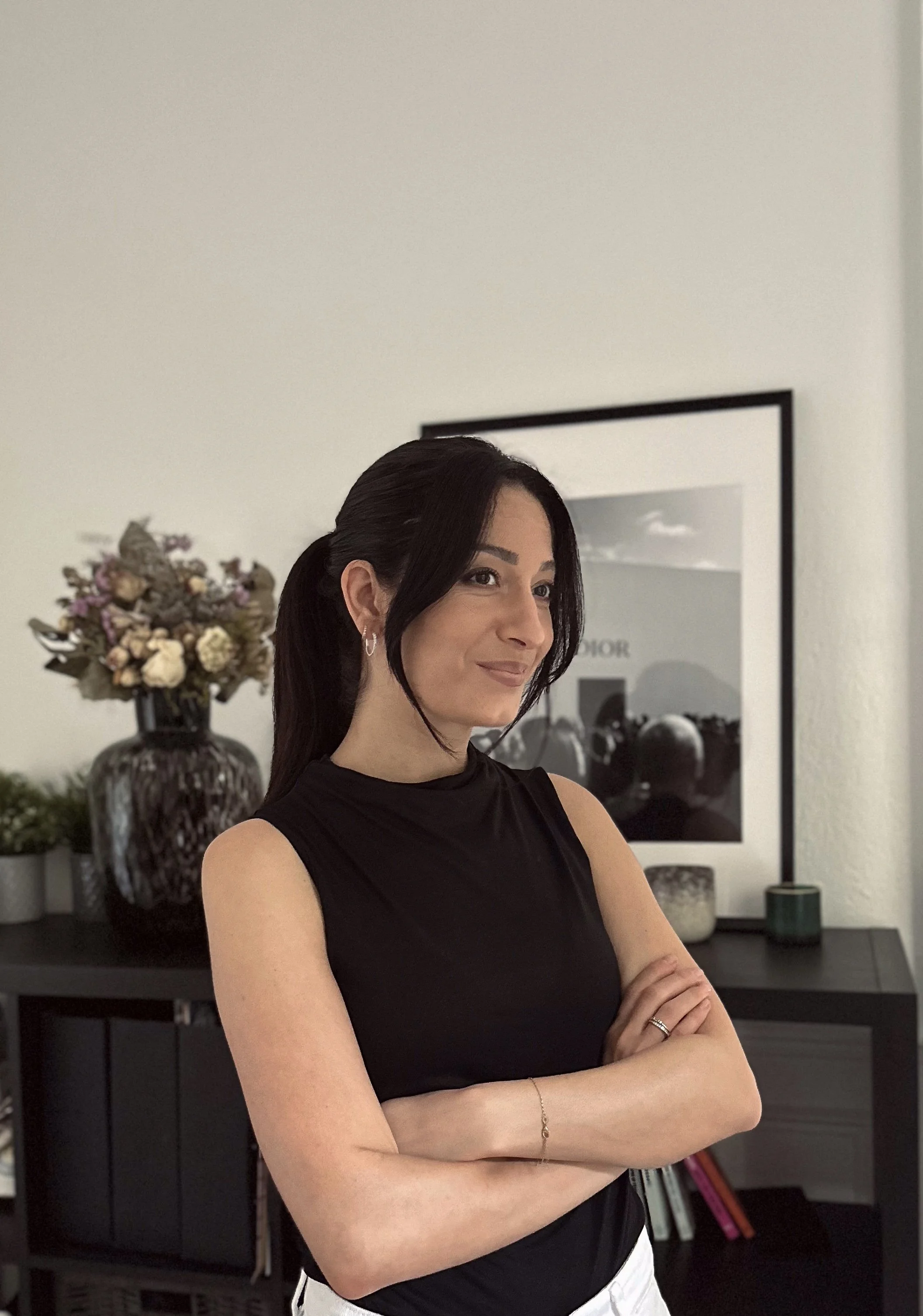 Une femme aux cheveux noirs, portant un haut sans manches noir, se tient dans une pièce moderne avec un meuble noir, un tableau noir et blanc au mur, une grande vase avec des fleurs séchées et quelques livres en arrière-plan.