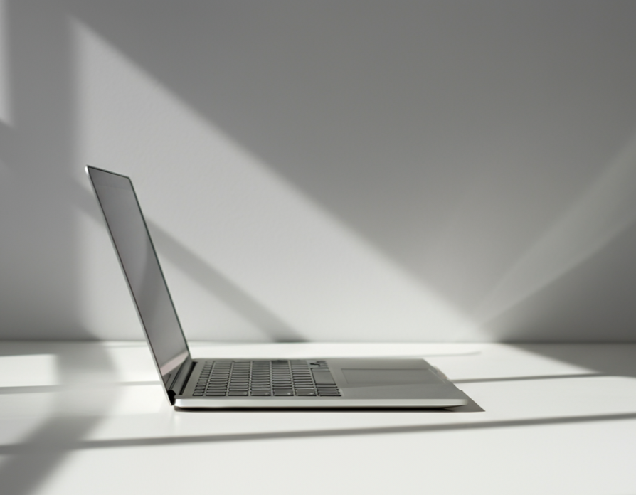 Un ordinateur portable placé sur une surface blanche avec un fond blanc, avec la lumière naturelle créant des ombres sur la surface.