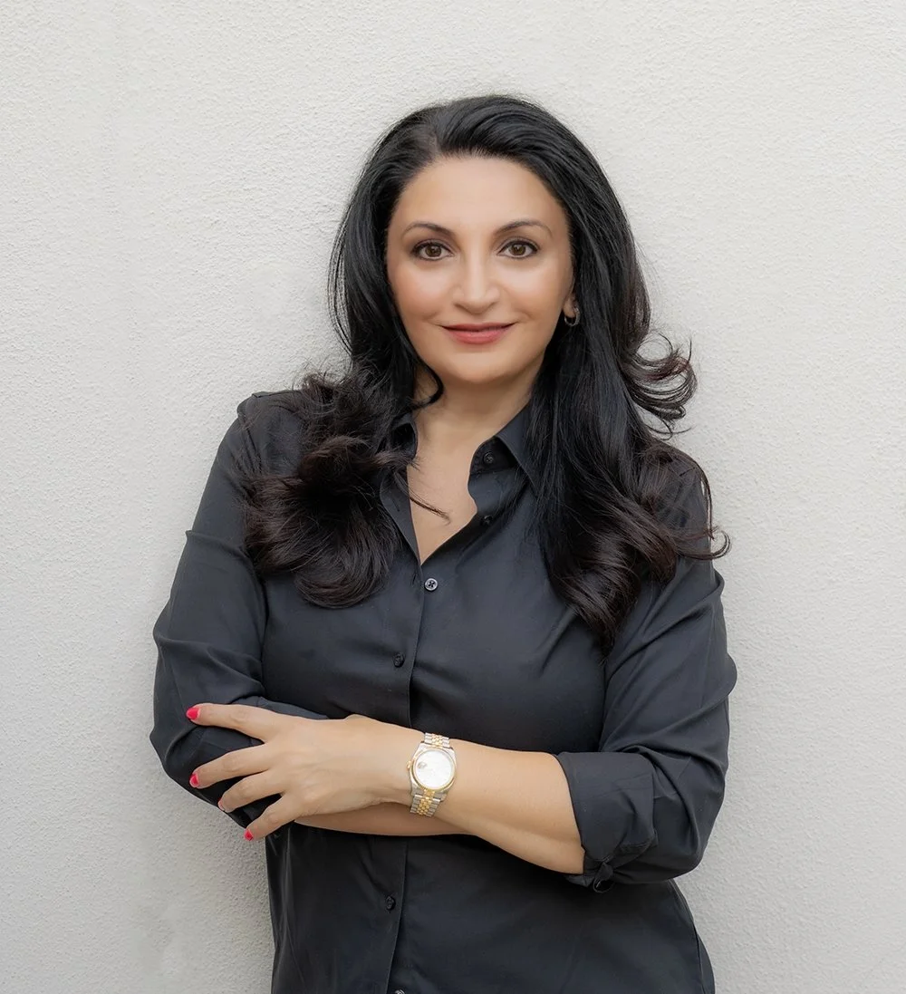A woman with long, dark, wavy hair, wearing a black shirt with sleeves rolled up, standing against a plain light-colored wall.