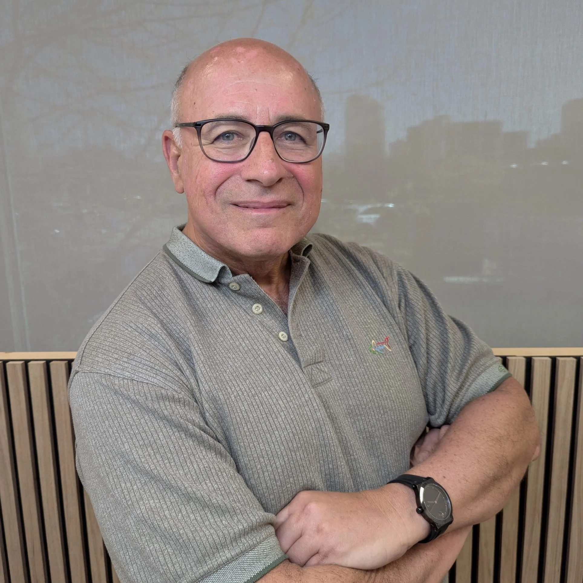 A middle-aged man with light skin, glasses, and a shaved head, smiling with his arms crossed, wearing a gray polo shirt and a wristwatch, standing in front of a light-colored wall with wood paneling at the bottom.