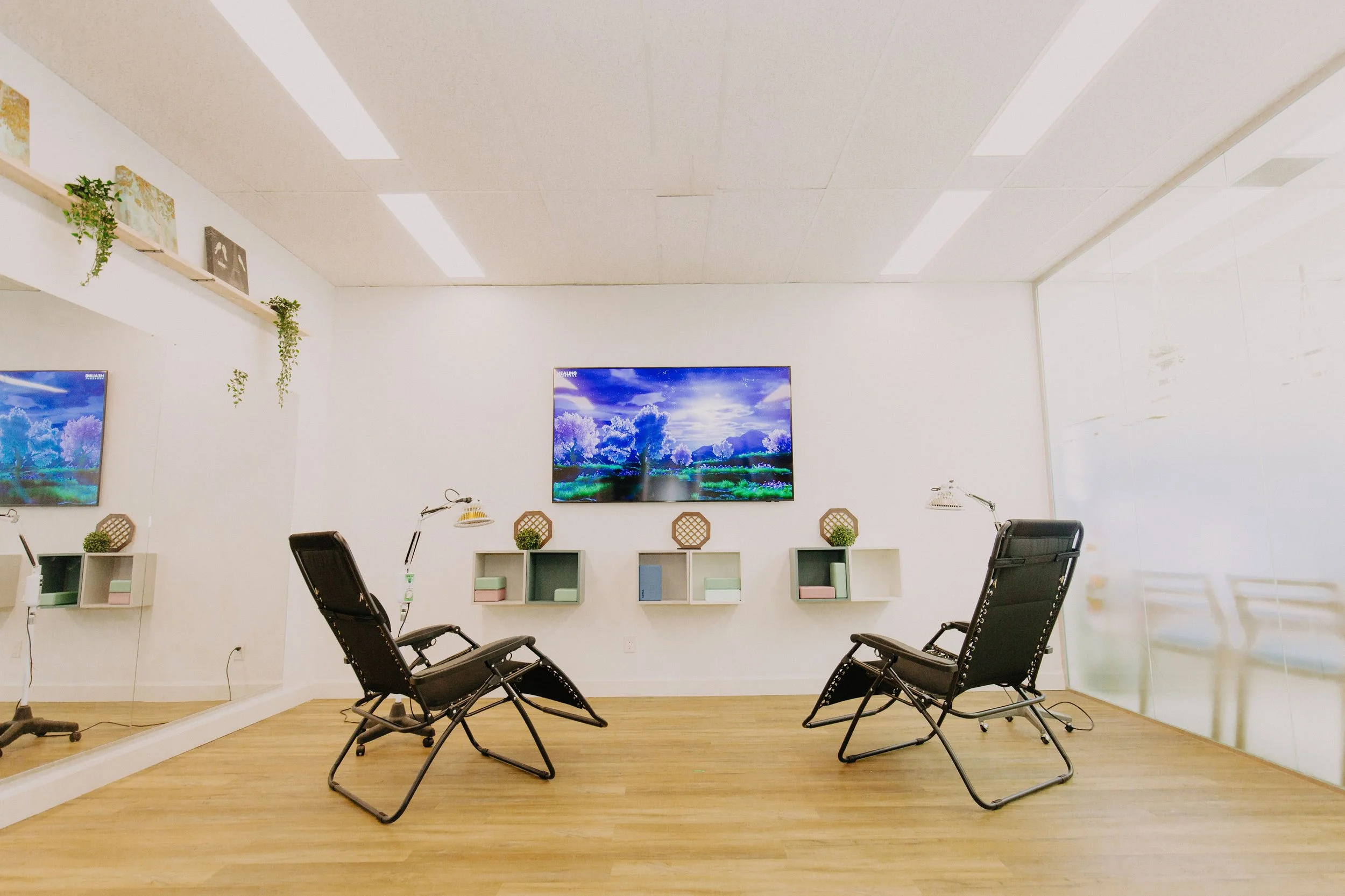 A modern meeting room with five black office chairs arranged around a small wooden table, with two wall-mounted TVs displaying nature scenes, and decorative shelves with plants and books on a white wall.