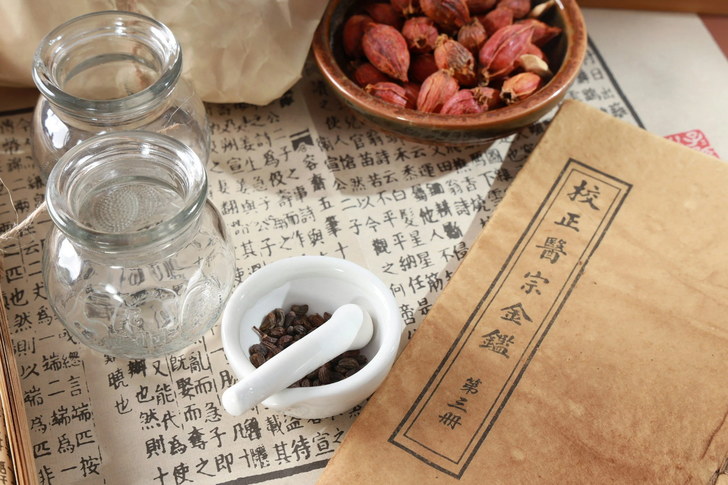 Traditional Chinese medicine ingredients and tools, including glass jars, a bowl of dried herbs, a small mortar and pestle with dried herbs, a paper with Chinese characters, and a brown paper envelope with Chinese writing.