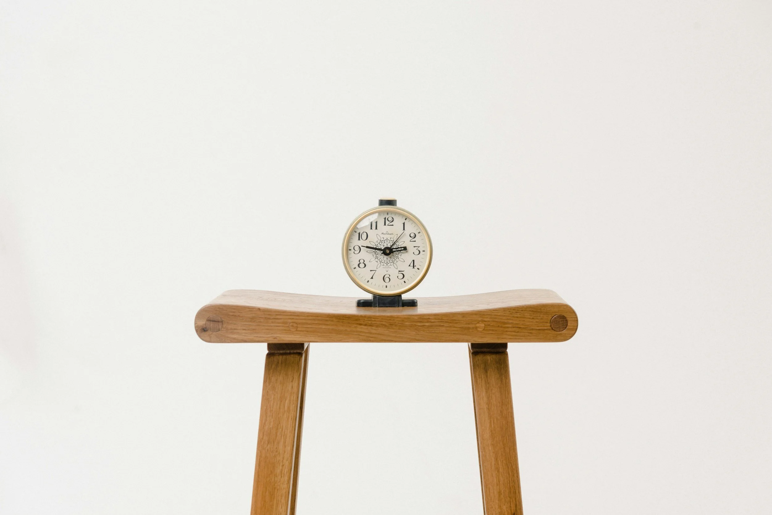 A small round clock with a white face and black numbers is placed on a wooden stool with simple legs against a plain white background.
