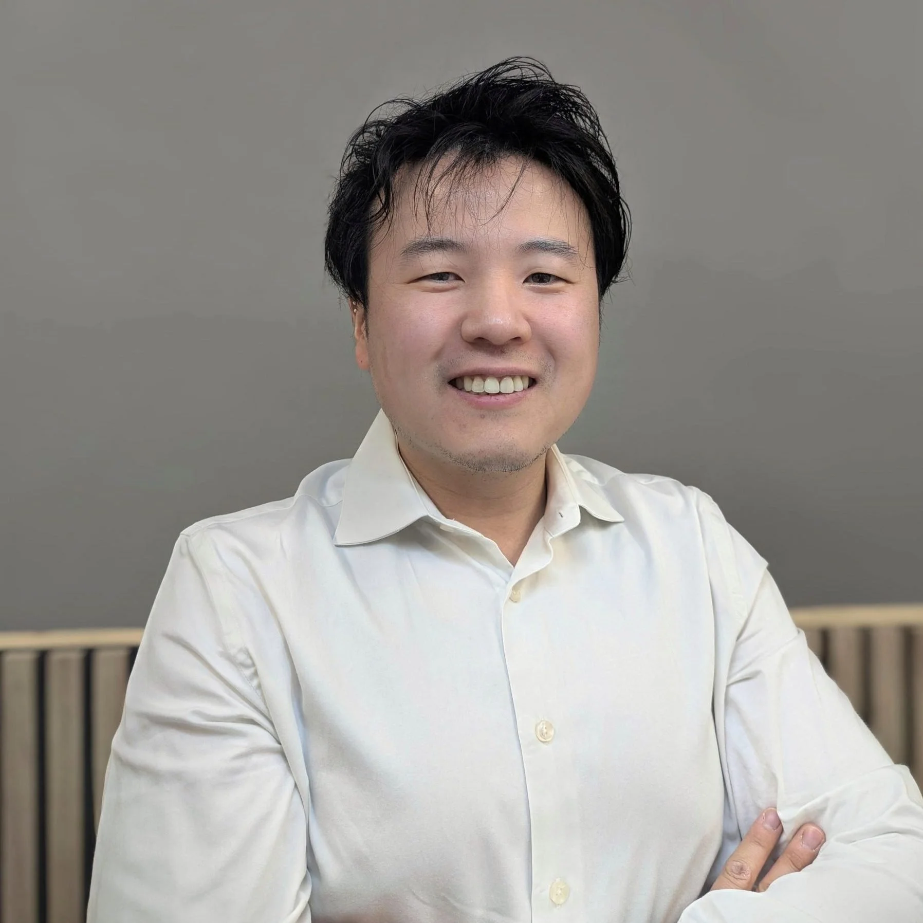 A smiling man with black, slightly messy hair, wearing a white dress shirt, sitting with arms crossed against a plain background.
