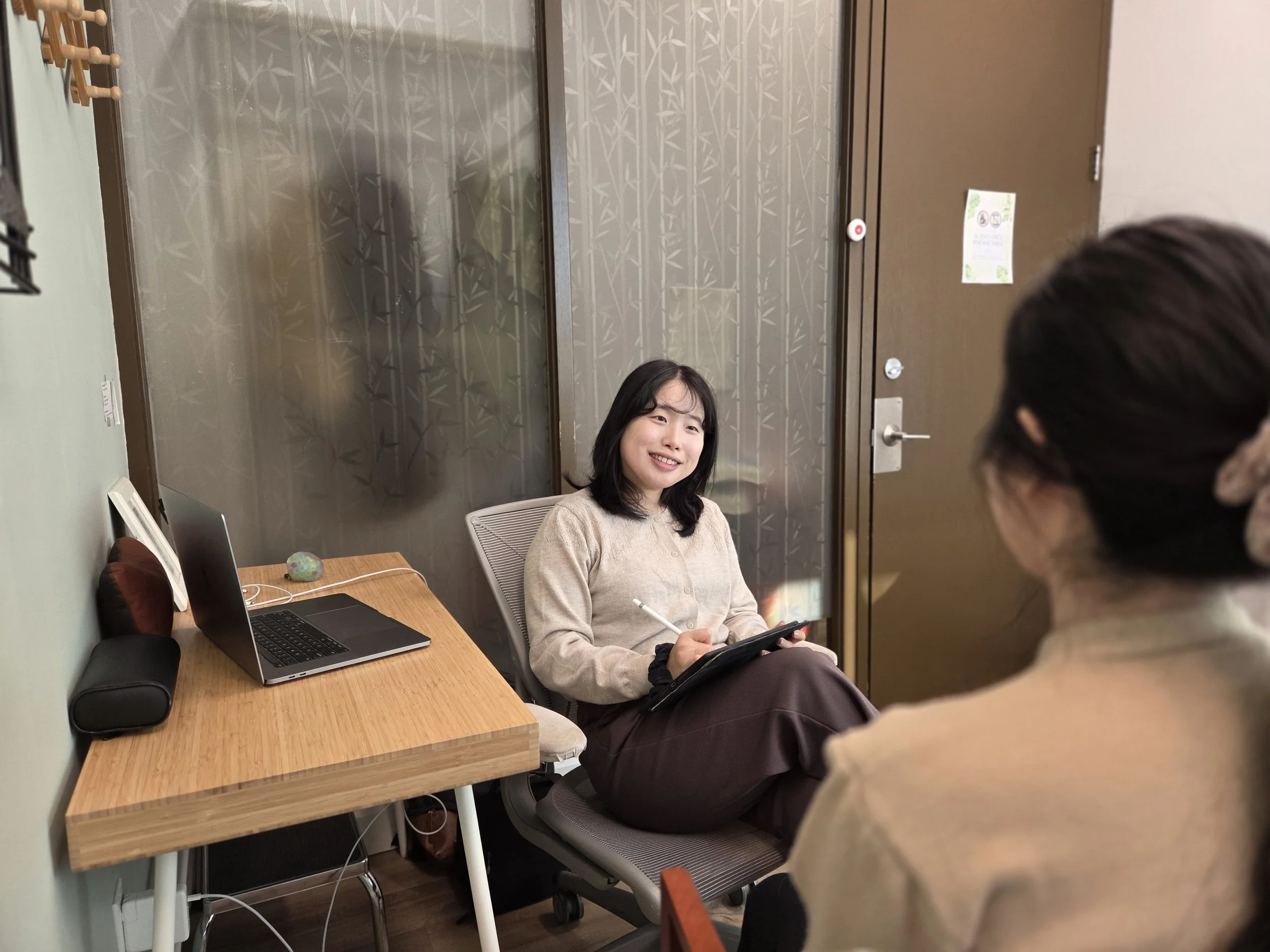 A woman with shoulder-length black hair and a beige sweater sits at a wooden desk smiling, holding a tablet. A man with dark hair is in the foreground, facing her. The desk has a laptop, a speaker, and some cables. The background features a brown door, a wall with bamboo-patterned wallpaper, and a coat hook with some hooks and a paper sign.
