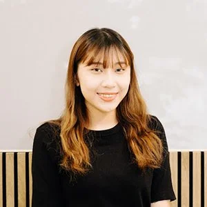 A young woman with long light brown hair and bangs, smiling, wearing a black shirt, standing in front of a white wall with a wooden slat pattern.