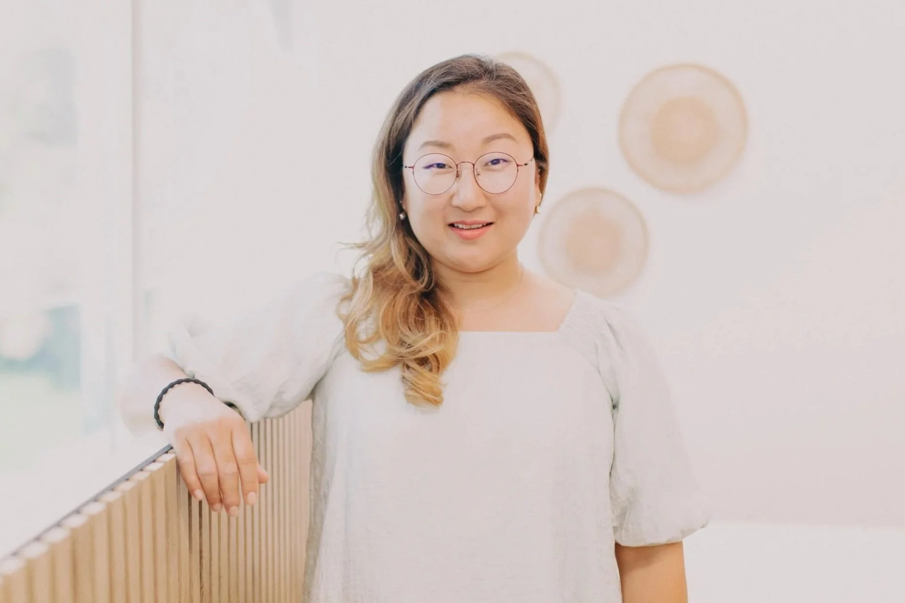 A woman with glasses and shoulder-length hair, wearing a white dress, smiling and leaning on a wooden railing in an indoor setting with white walls and circular decorations.