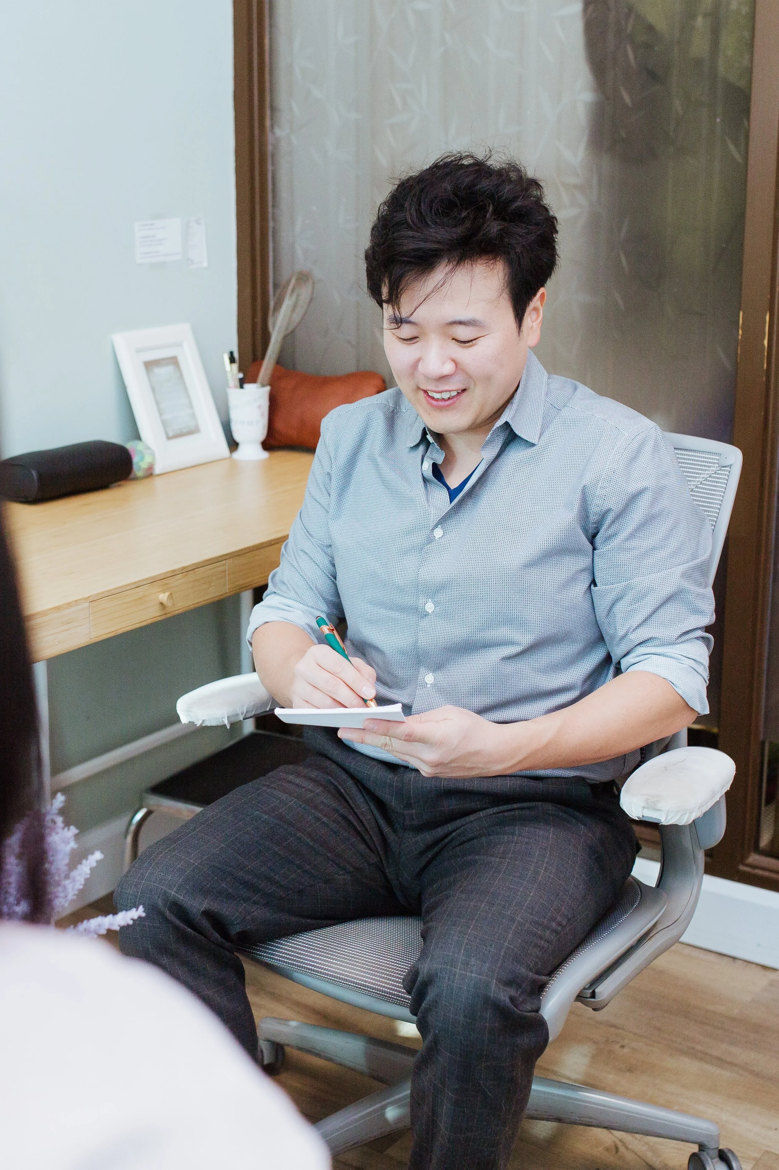 A man with dark hair, wearing a light gray button-up shirt and dark pants, sitting on a gray office chair, smiling while writing on a notepad. In the background, there is a wooden desk with various items on it, including a picture frame, a cup with pens, and a black case. The room has a glass door with a frosted pattern and appears to be a home office or professional setting.