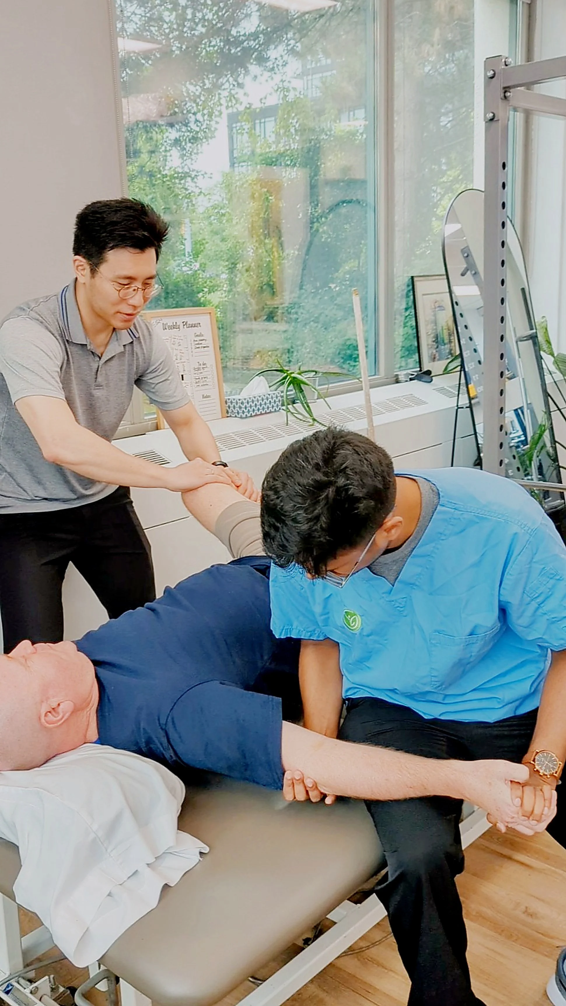 A man is receiving a medical procedure or assistance from two healthcare workers, one of whom is holding his arm, in a room with large windows and medical equipment.