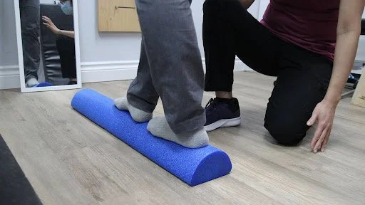 A person standing on a blue foam roller on the floor while a trainer or therapist kneels beside them, providing support or guidance.
