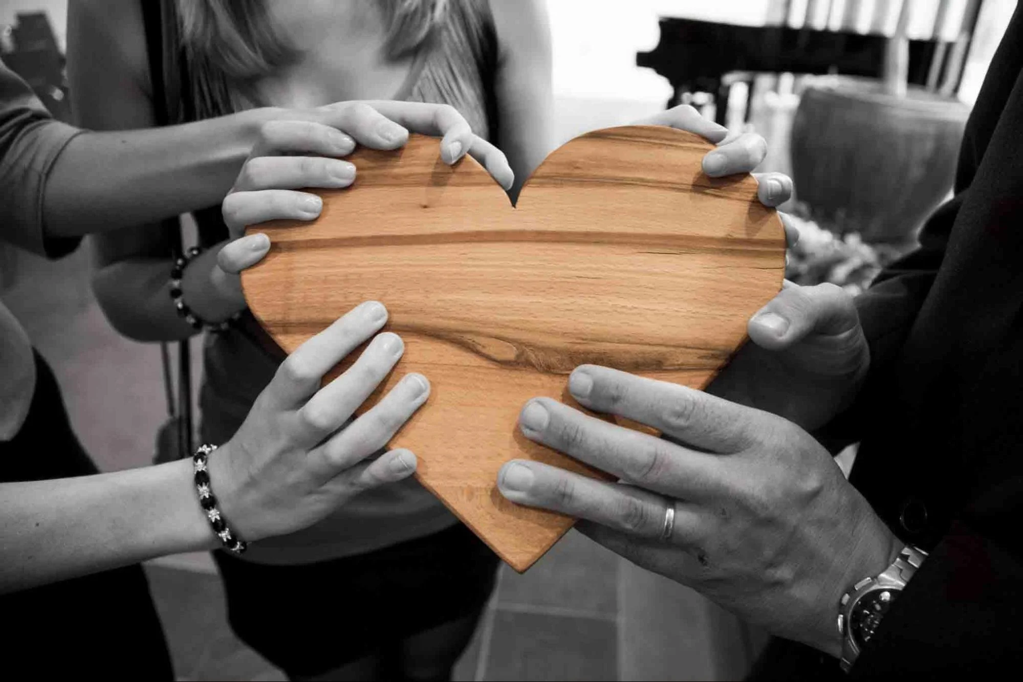 Multiple people's hands holding a wooden heart-shaped object together in black and white, with the central part of the image in color, showing the natural wood grain of the heart.