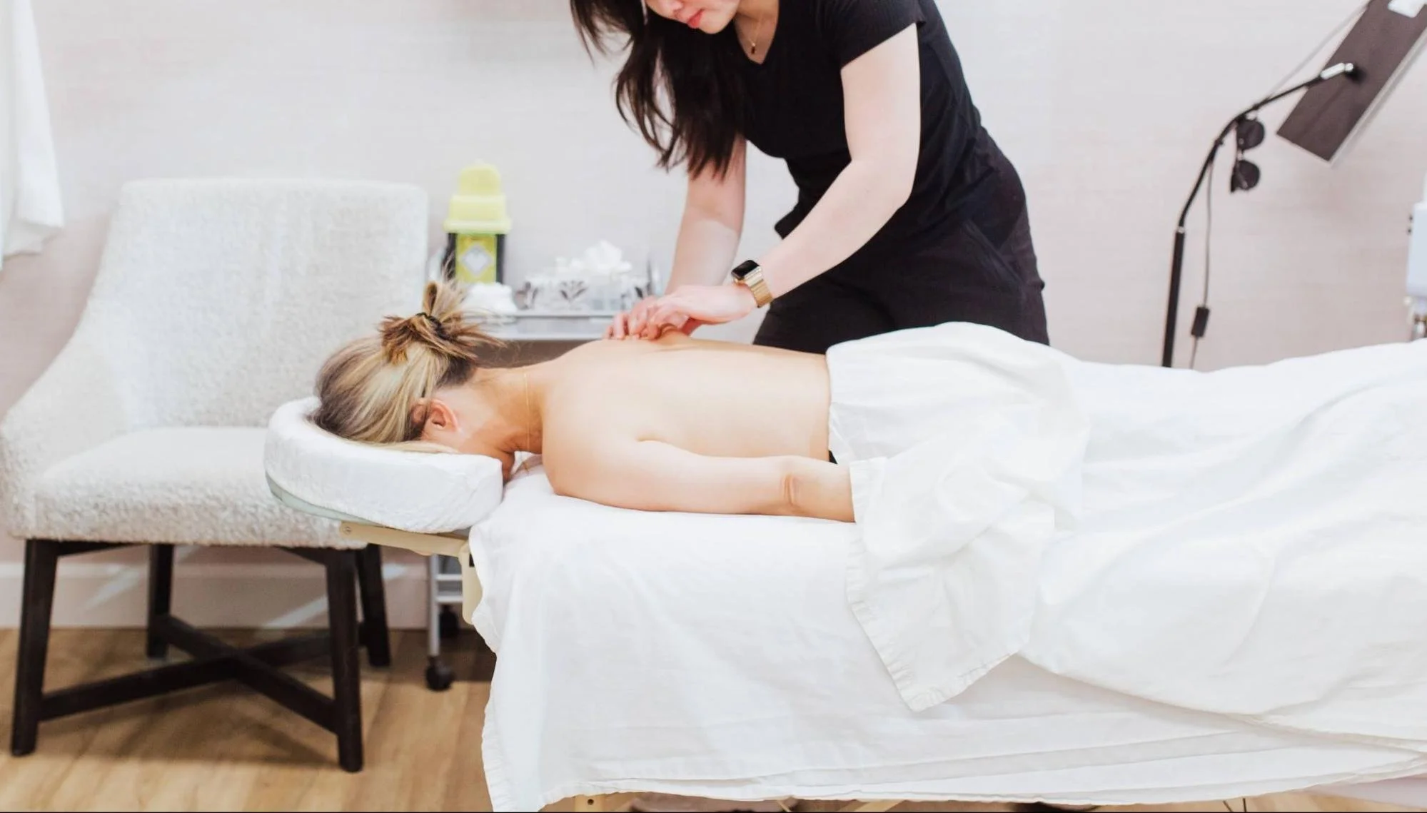 A woman receiving a massage in a modern treatment room with a couch, a table with supplies, and professional lighting.