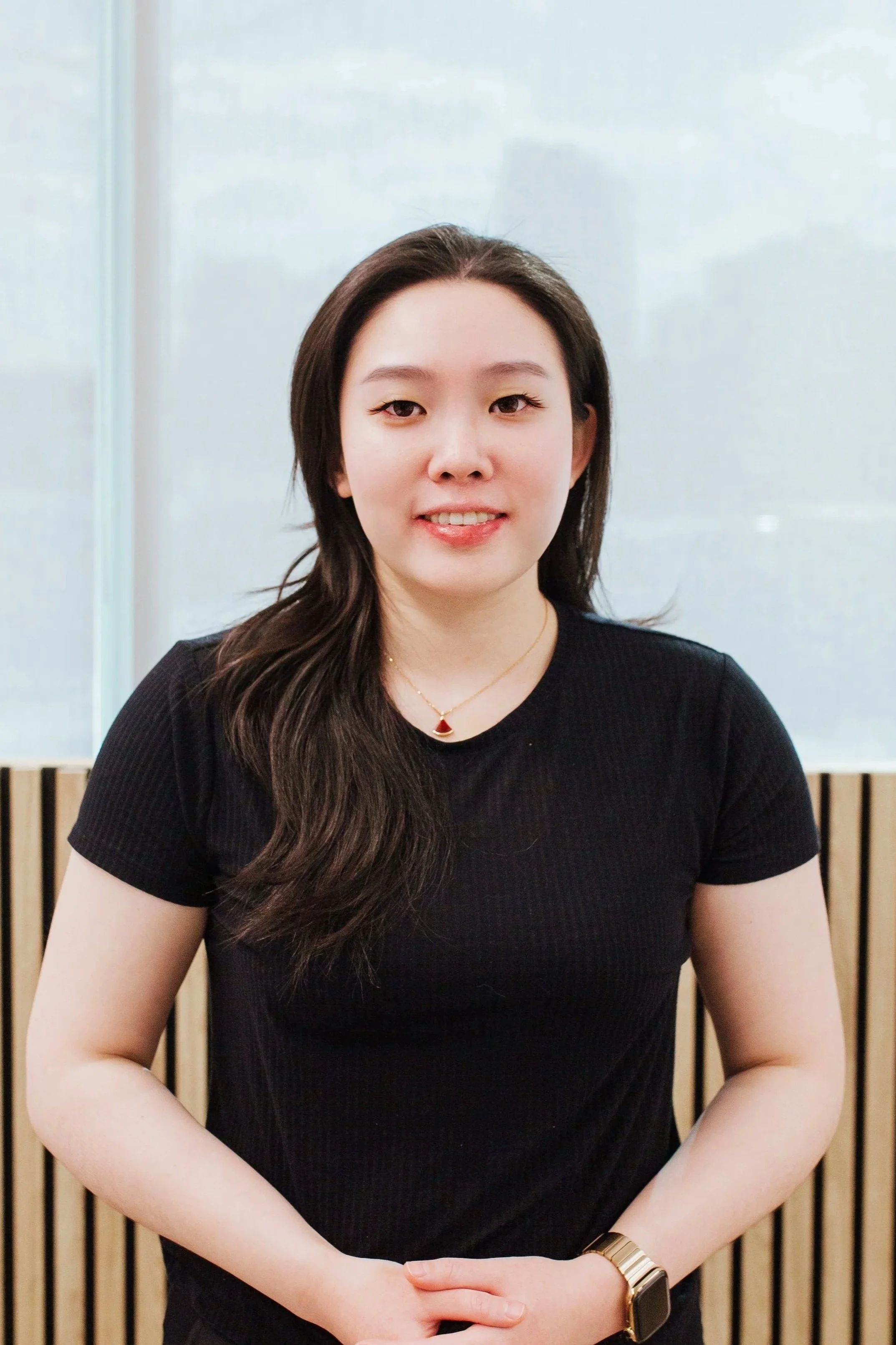 A young woman with long, dark wavy hair is standing in front of a background with vertical wooden slats and a frosted glass panel. She is wearing a black, short-sleeved top, a gold necklace with a red pendant, and a gold watch on her left wrist. She has a slight smile and is looking directly at the camera.