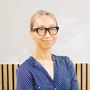 A woman with blonde hair, glasses, and a blue polka dot blouse standing in front of a plain background with wooden paneling.