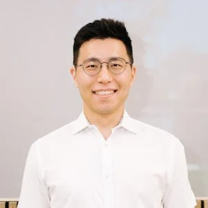 A young man with glasses and short dark hair, smiling, wearing a white shirt, standing against a neutral background.