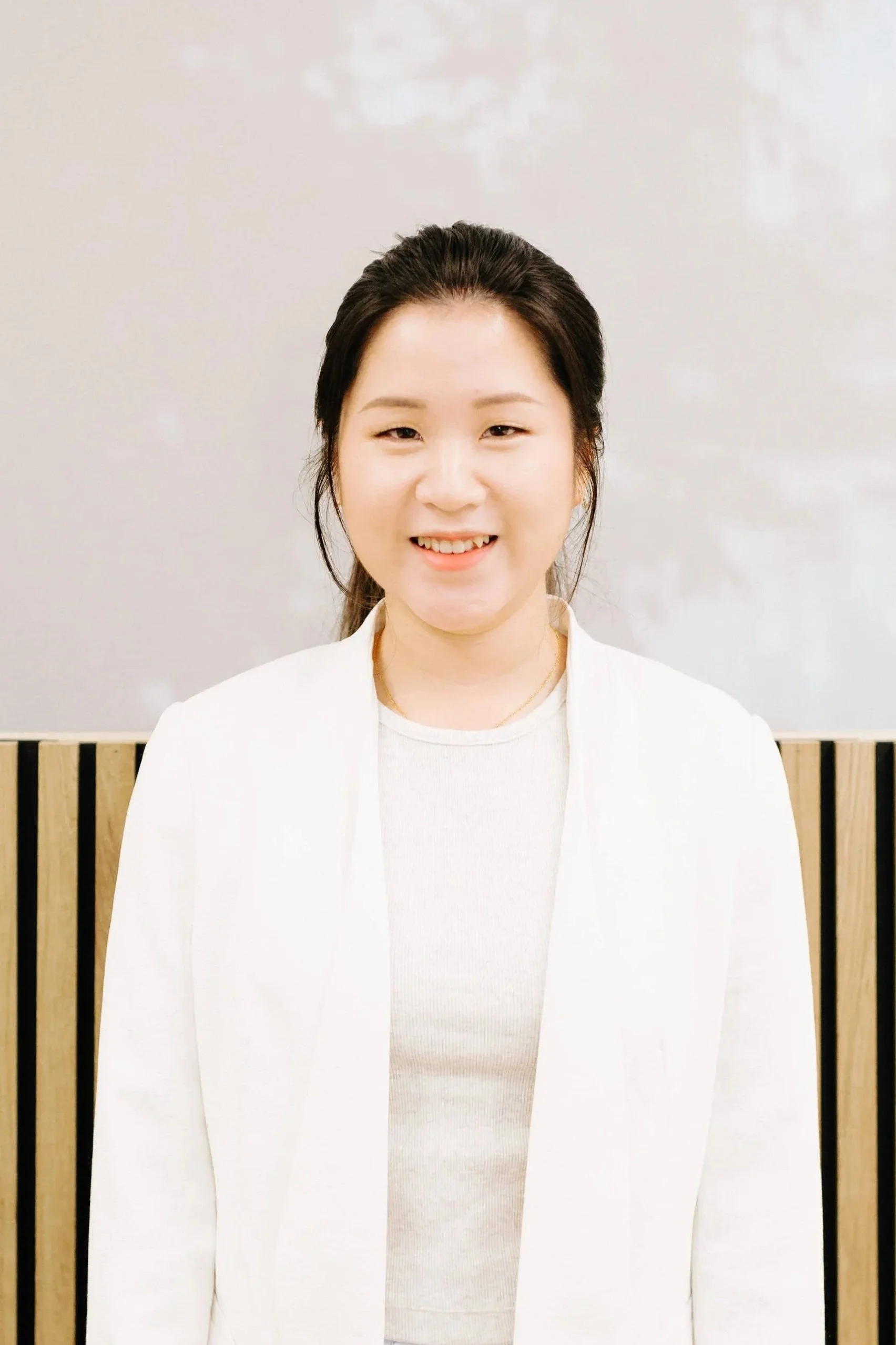A young woman with dark hair in a low ponytail, smiling and wearing a white blazer over a white top, standing in front of a light-colored wall with wooden slats at the bottom.