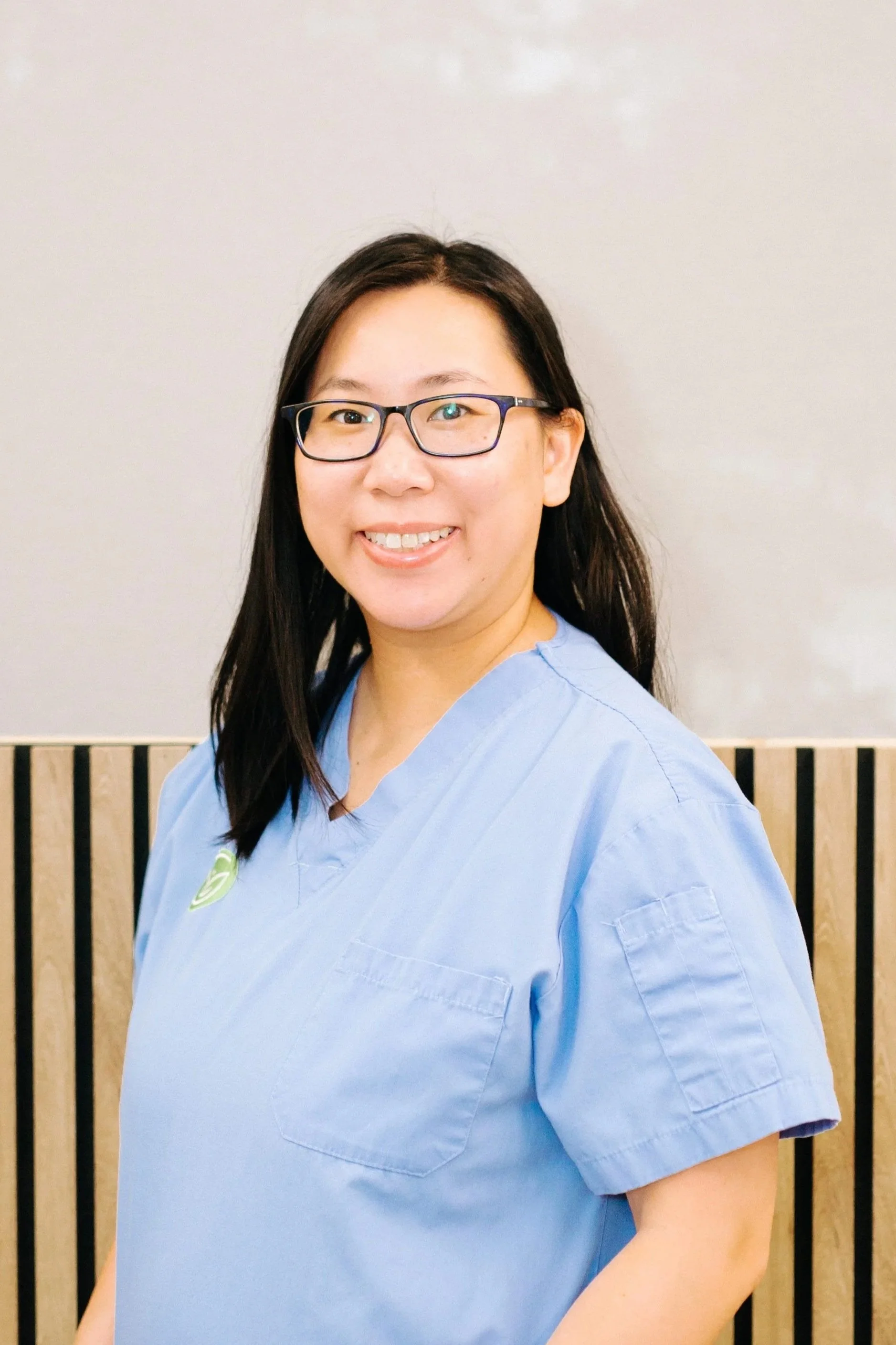 Close-up of a smiling woman with long dark hair and glasses wearing light blue medical scrubs, standing in front of a wooden slat wall.