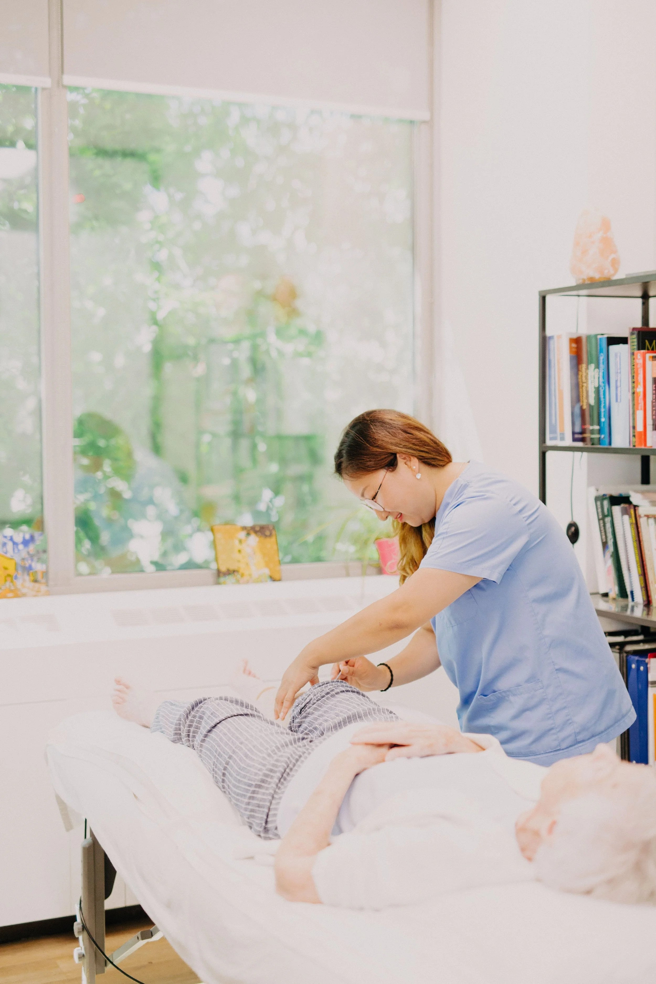 A healthcare professional in light blue scrubs attending to a patient lying on a hospital bed in a bright room with large windows and a bookshelf.
