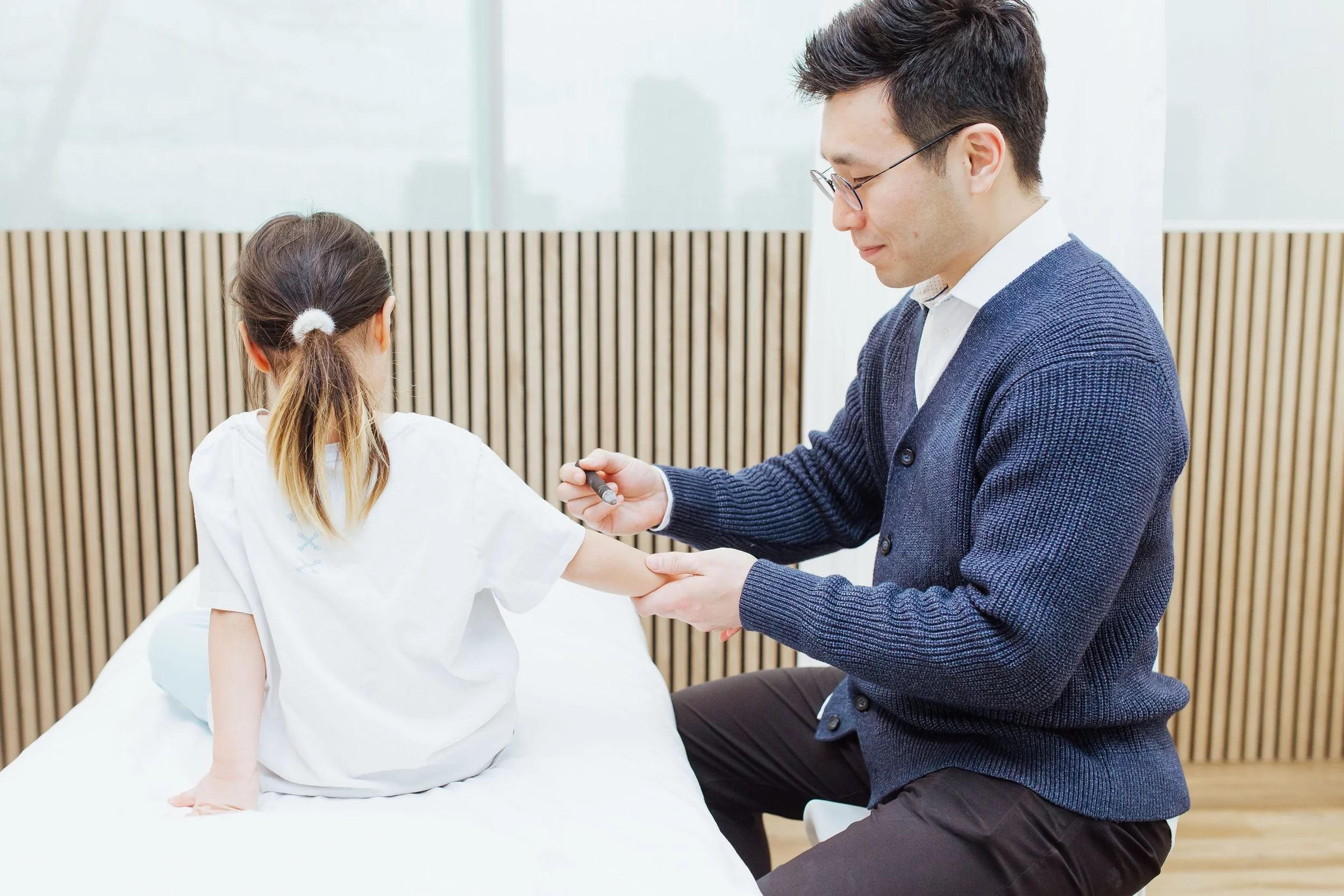 A man in glasses and a navy blue sweater helping a young girl sit up in a medical or educational setting, with a background of wooden paneling and large windows.