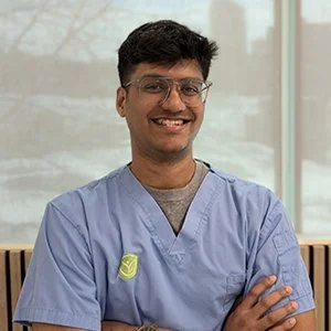 Young man in medical scrubs smiling, arms crossed, indoors with large windows in the background.