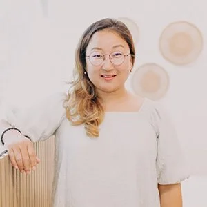 A woman with glasses and long dark hair smiling in front of a wooden wall panel.