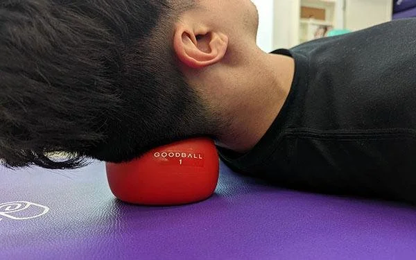 Person lying on a purple mat with their head resting on a red massage ball labeled "Goodball 1."
