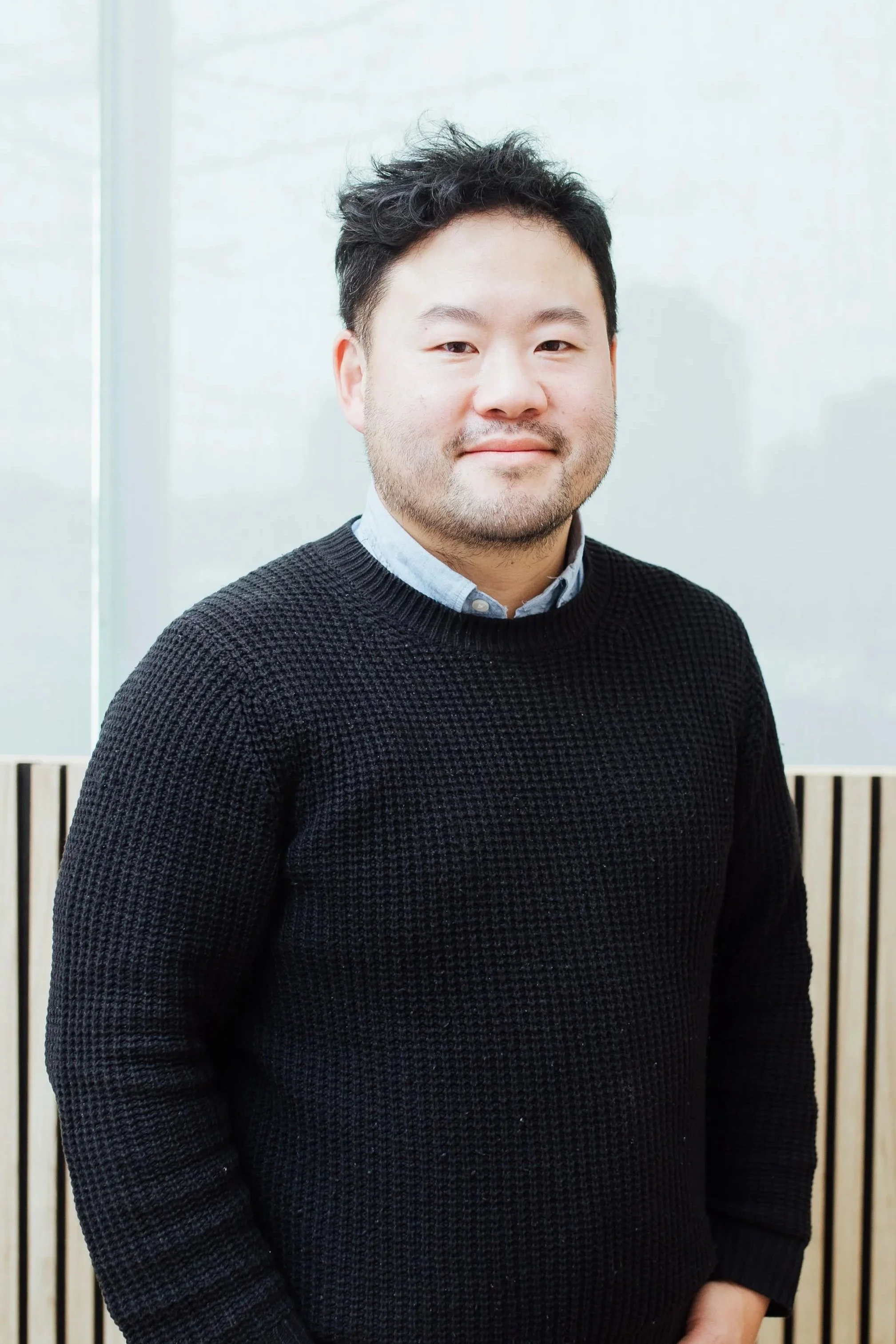 Portrait of a man with short black hair, light skin, and a beard, wearing a dark textured sweater over a light collared shirt, standing in front of a plain white background with wooden slats.