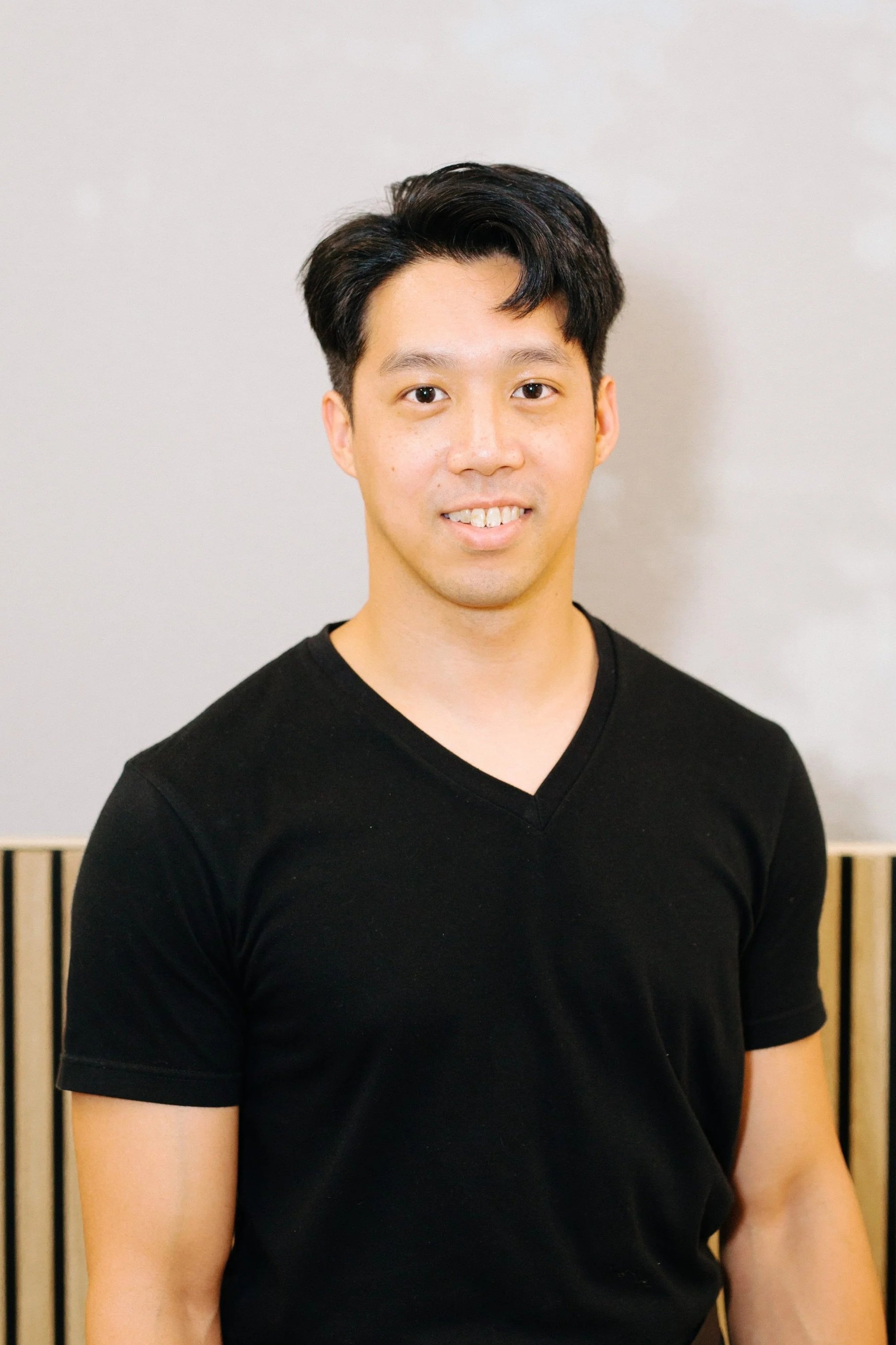 A young man with black hair and a black shirt smiling in front of a light-colored wall and wooden slats.