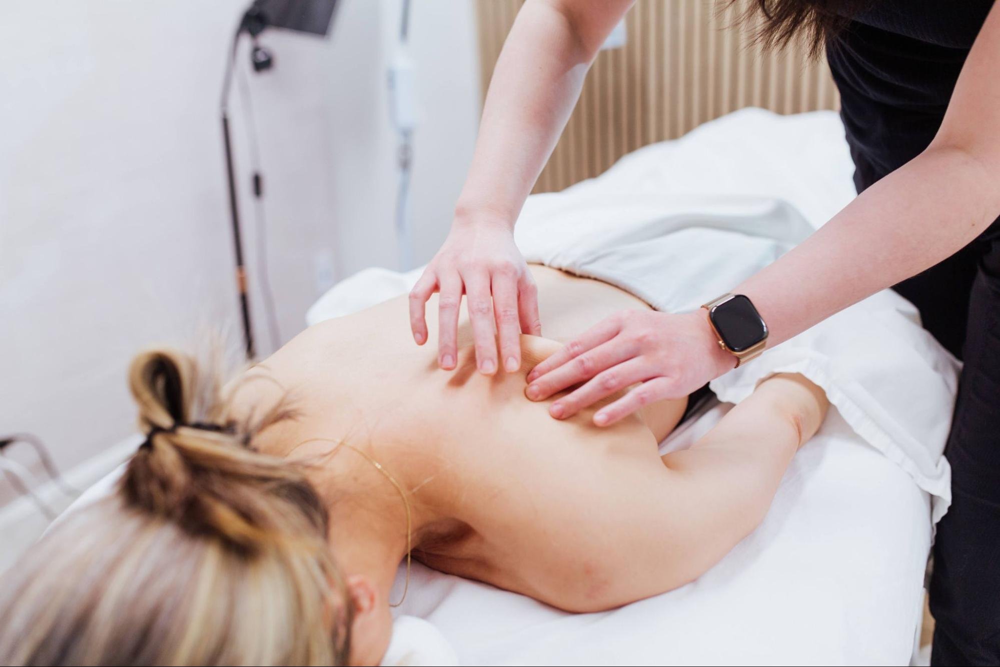 A person receiving a massage on their back from a massage therapist in a clinical setting. The therapist's hands are pressing on the person's back.