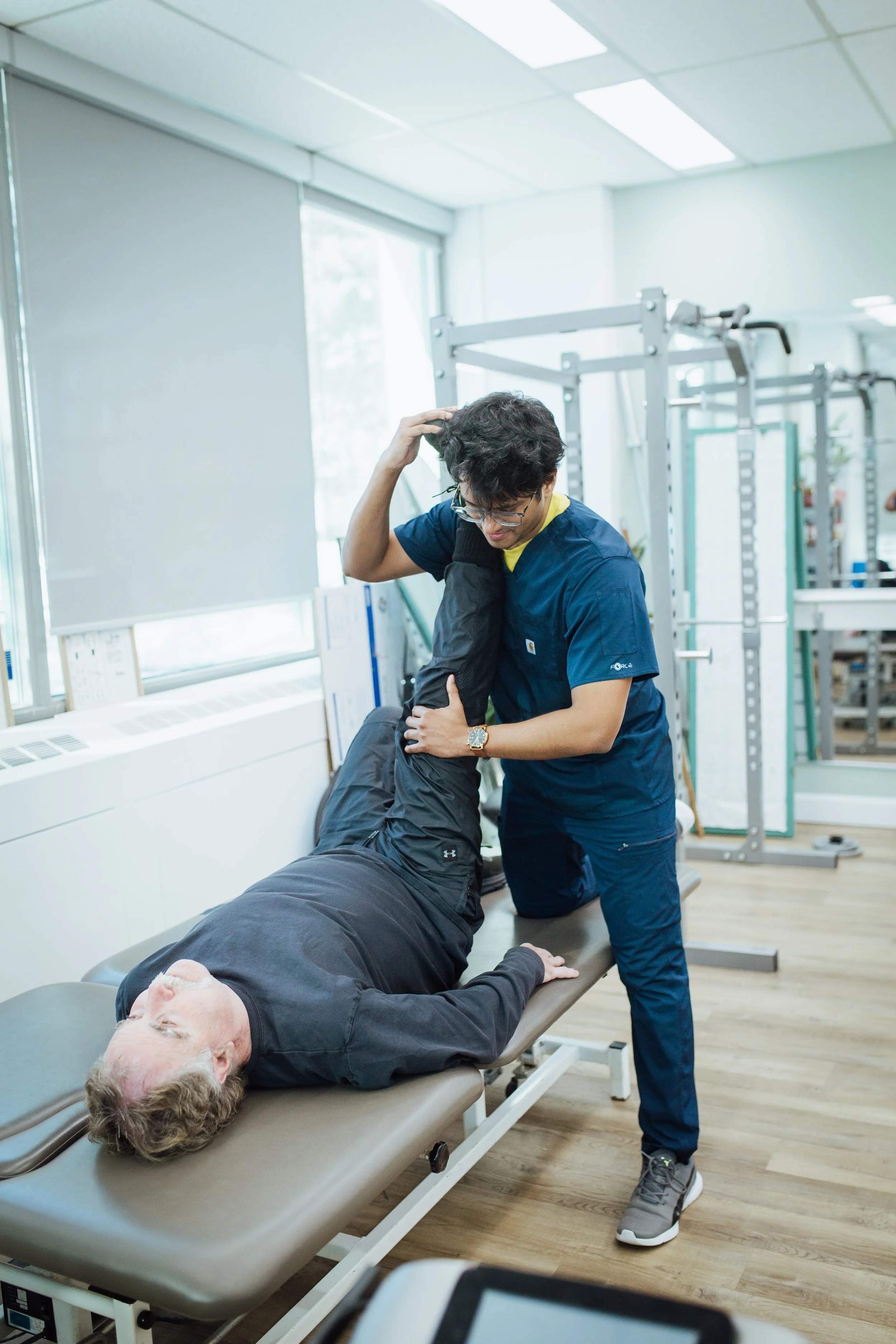 Physical therapist assisting an elderly man with a leg stretch exercise on a therapy table in a clinical setting.