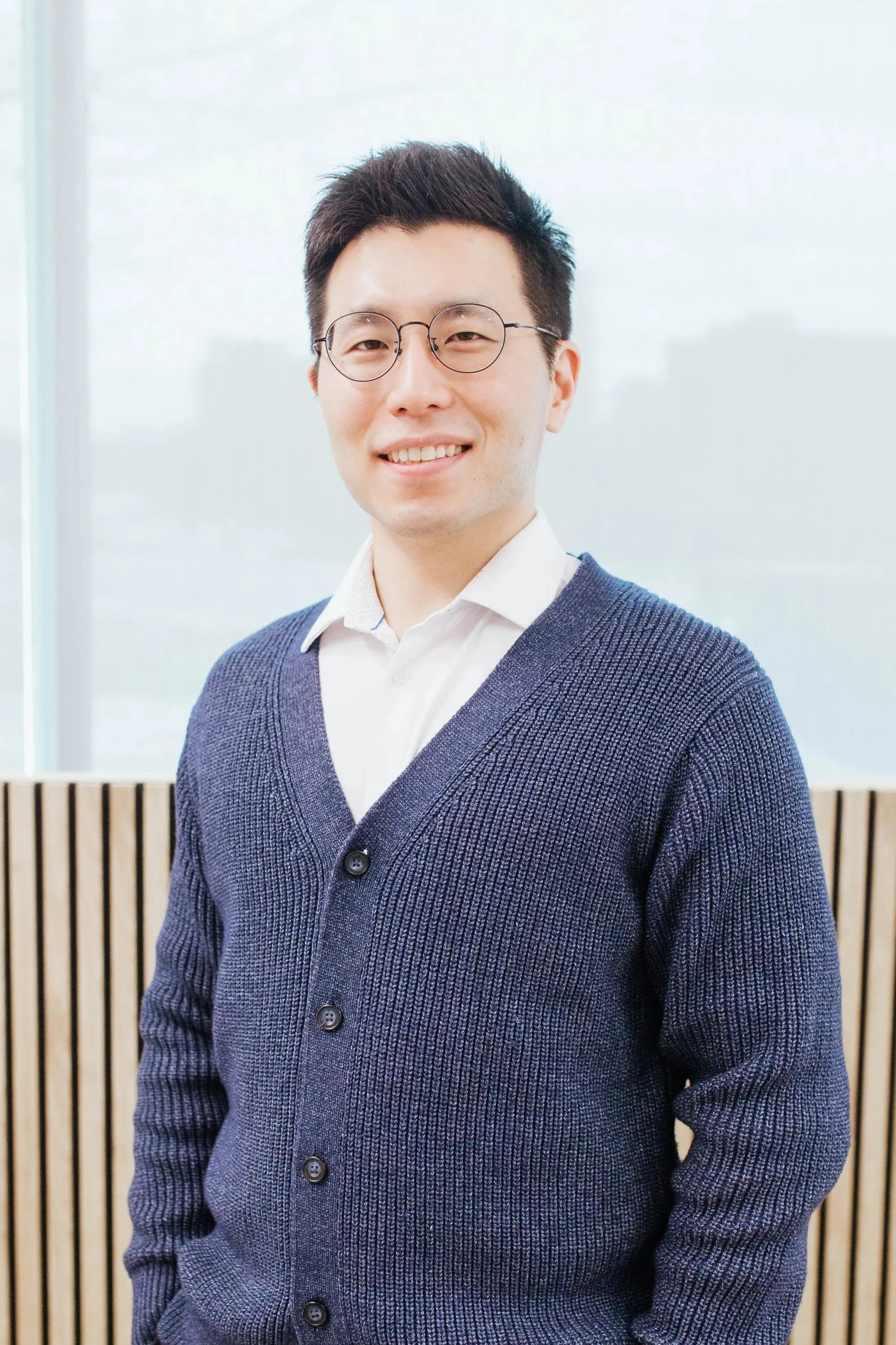 A young man with short dark hair, wearing round glasses, a white shirt, and a dark blue cardigan, smiling in front of a light-colored background with wooden slats and a frosted window.