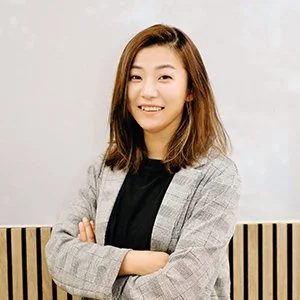 A young woman with shoulder-length brown hair smiling, wearing a plaid blazer over a black top, standing against a light background.