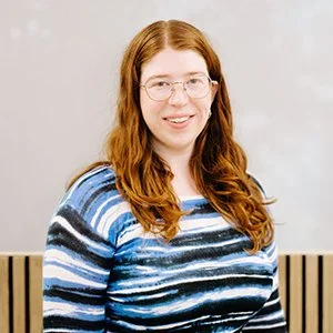 A woman with long red hair, glasses, and a blue striped sweater smiling in front of a light-colored wall.