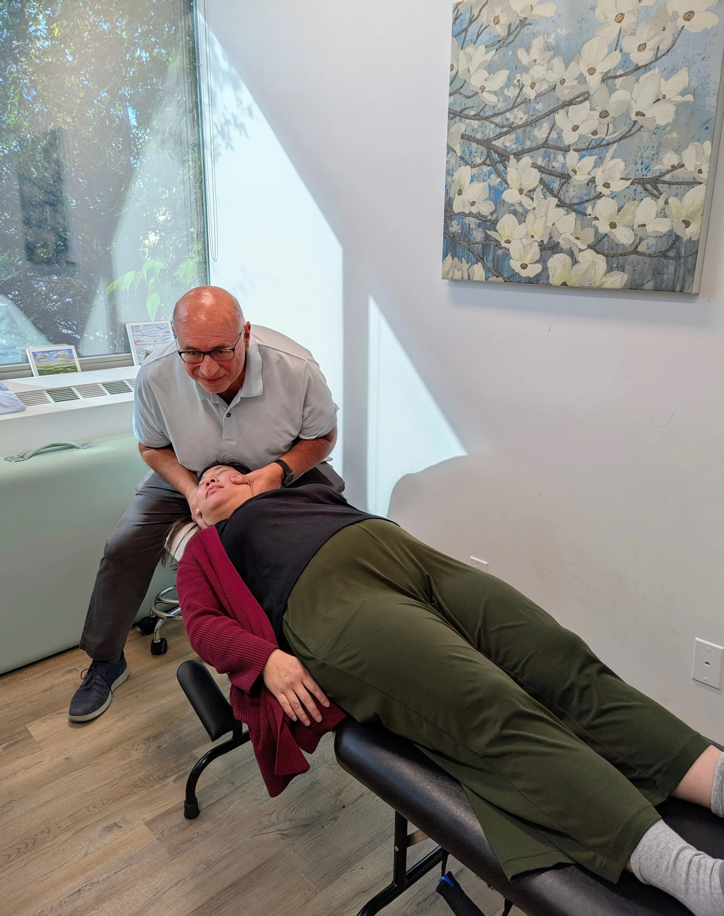 A man is lying face down on a massage table, receiving a neck massage from another man who is leaning over him, inside a room with a large window and a flower painting on the wall.
