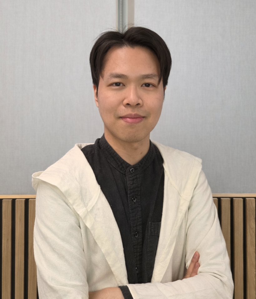 A young man with short black hair, wearing a black shirt and a cream-colored jacket, standing with his arms crossed in front of a light gray background with a wooden panel.