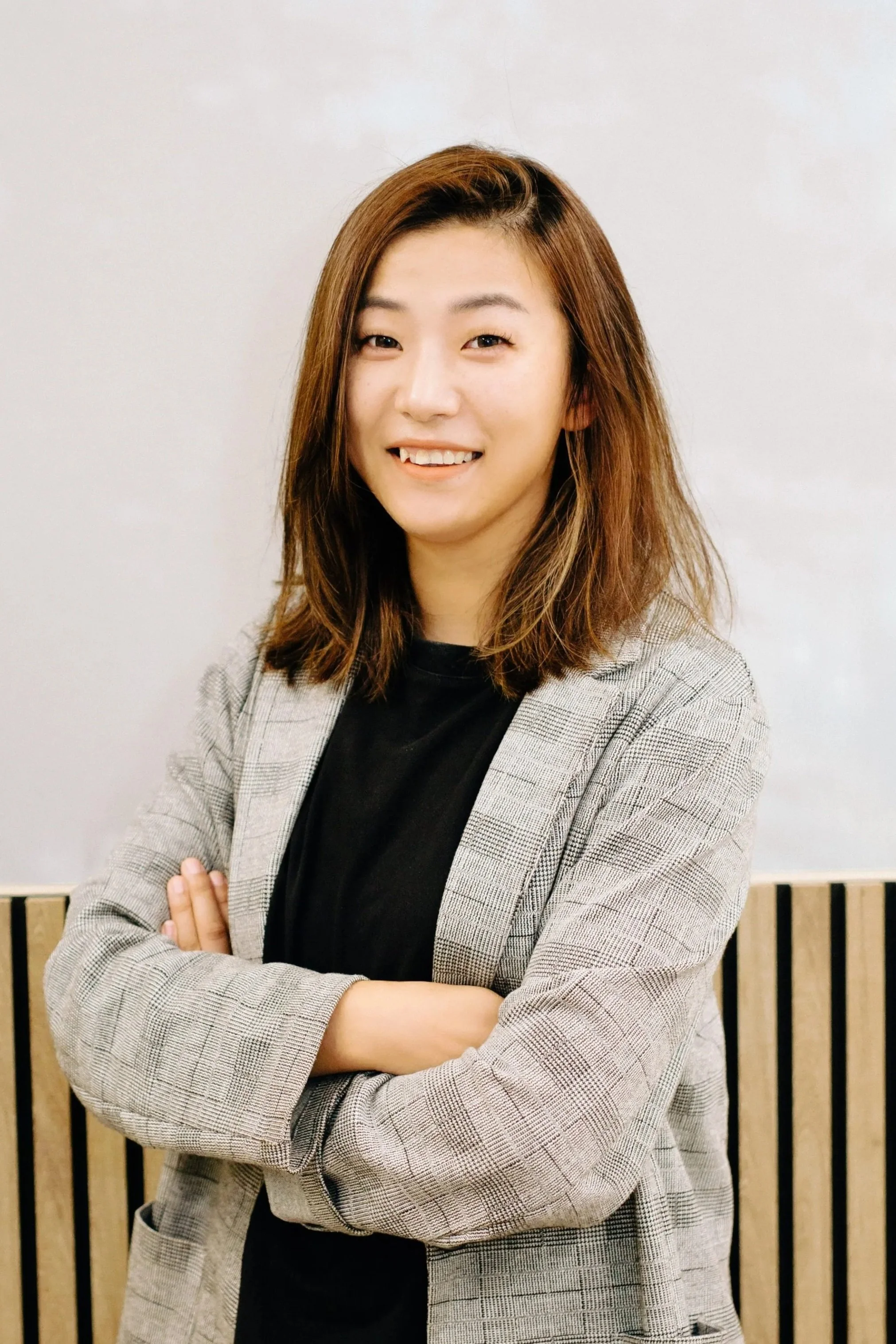 A young woman with shoulder-length brown hair, crossed arms, smiling, wearing a gray checked blazer over a black top, standing against a white wall with a wooden slat background.